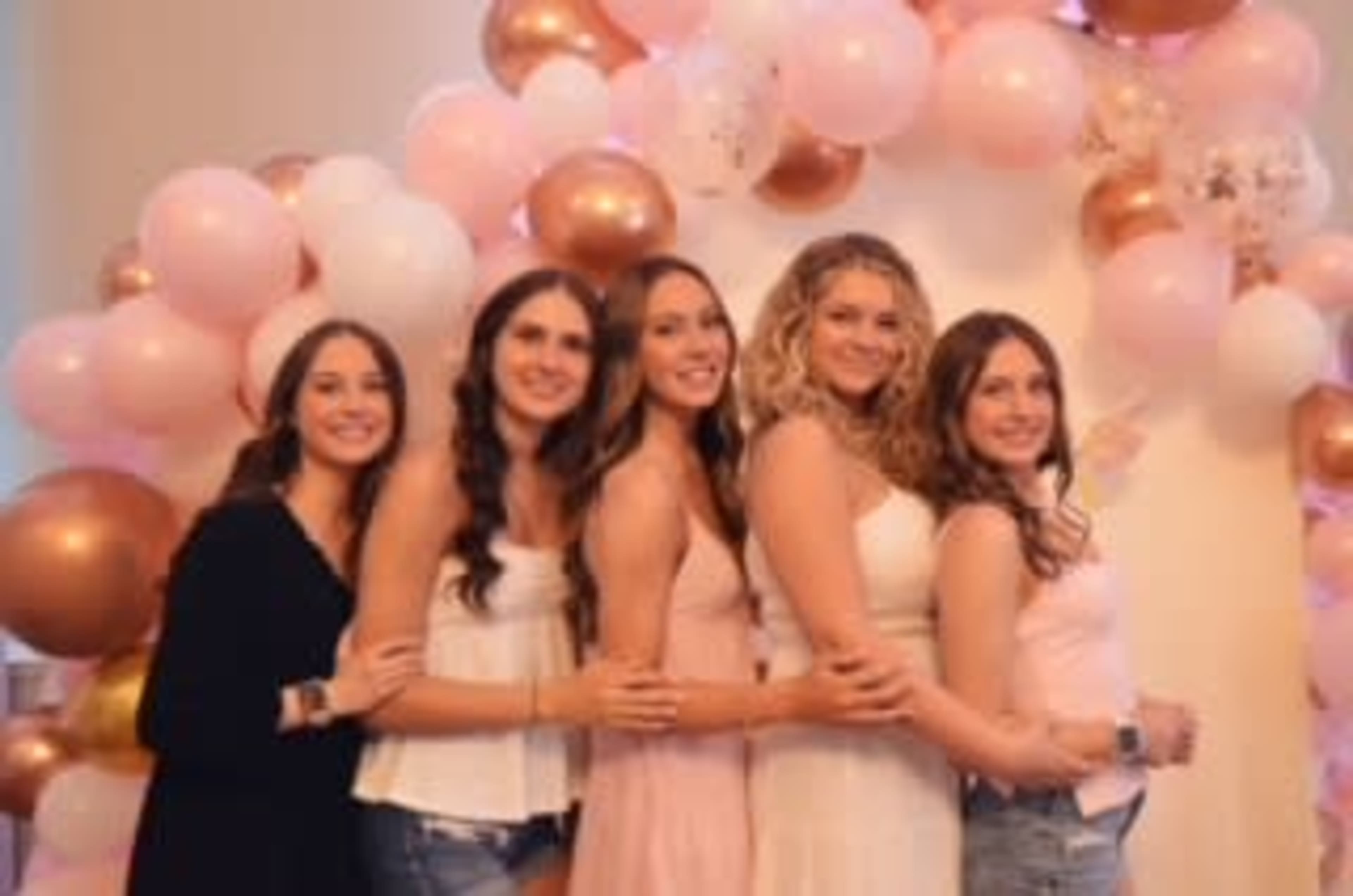 Five women stand together in front of a balloon arch made of pink and gold balloons, smiling at the camera.