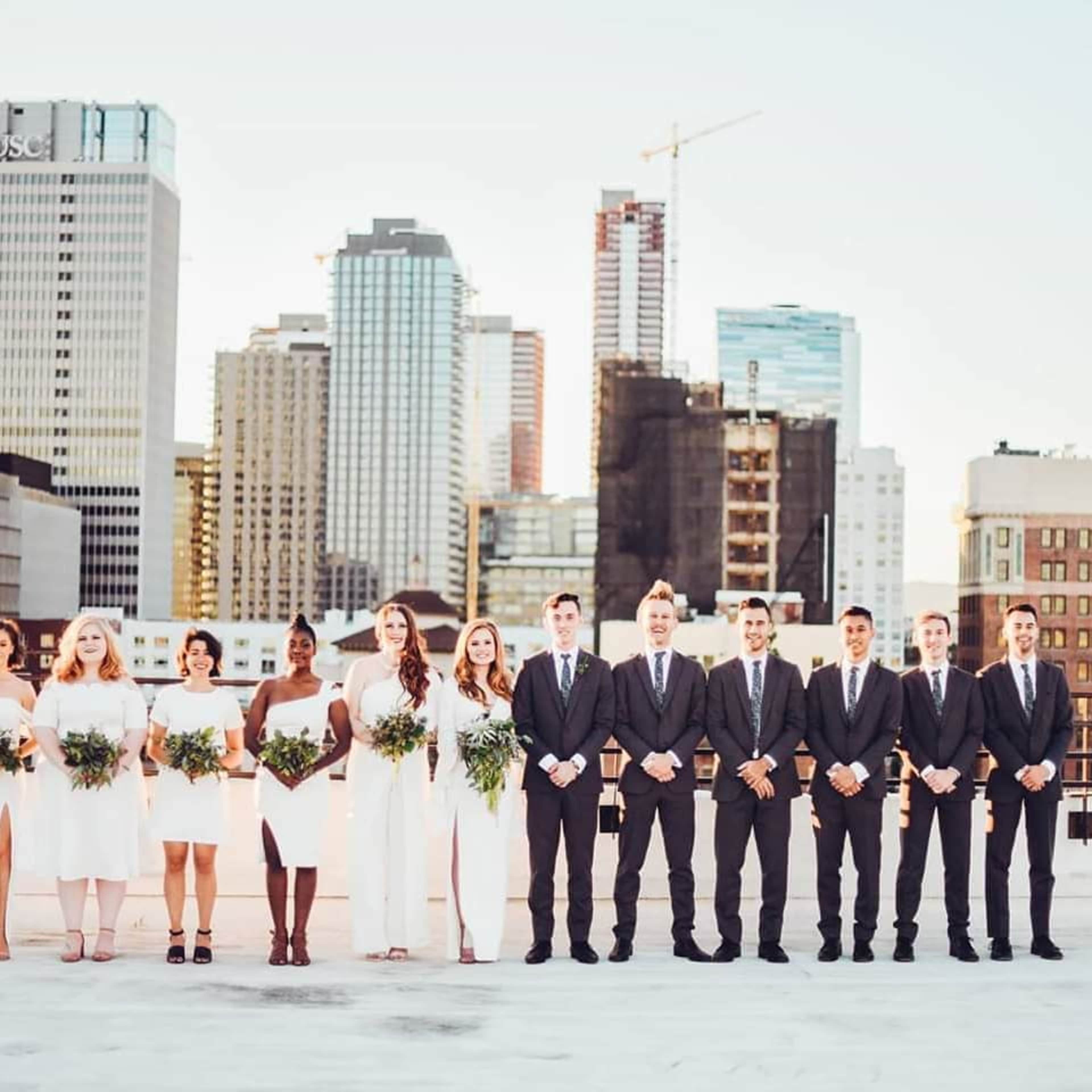 A wedding party stands in two rows on a rooftop with a city skyline in the background.