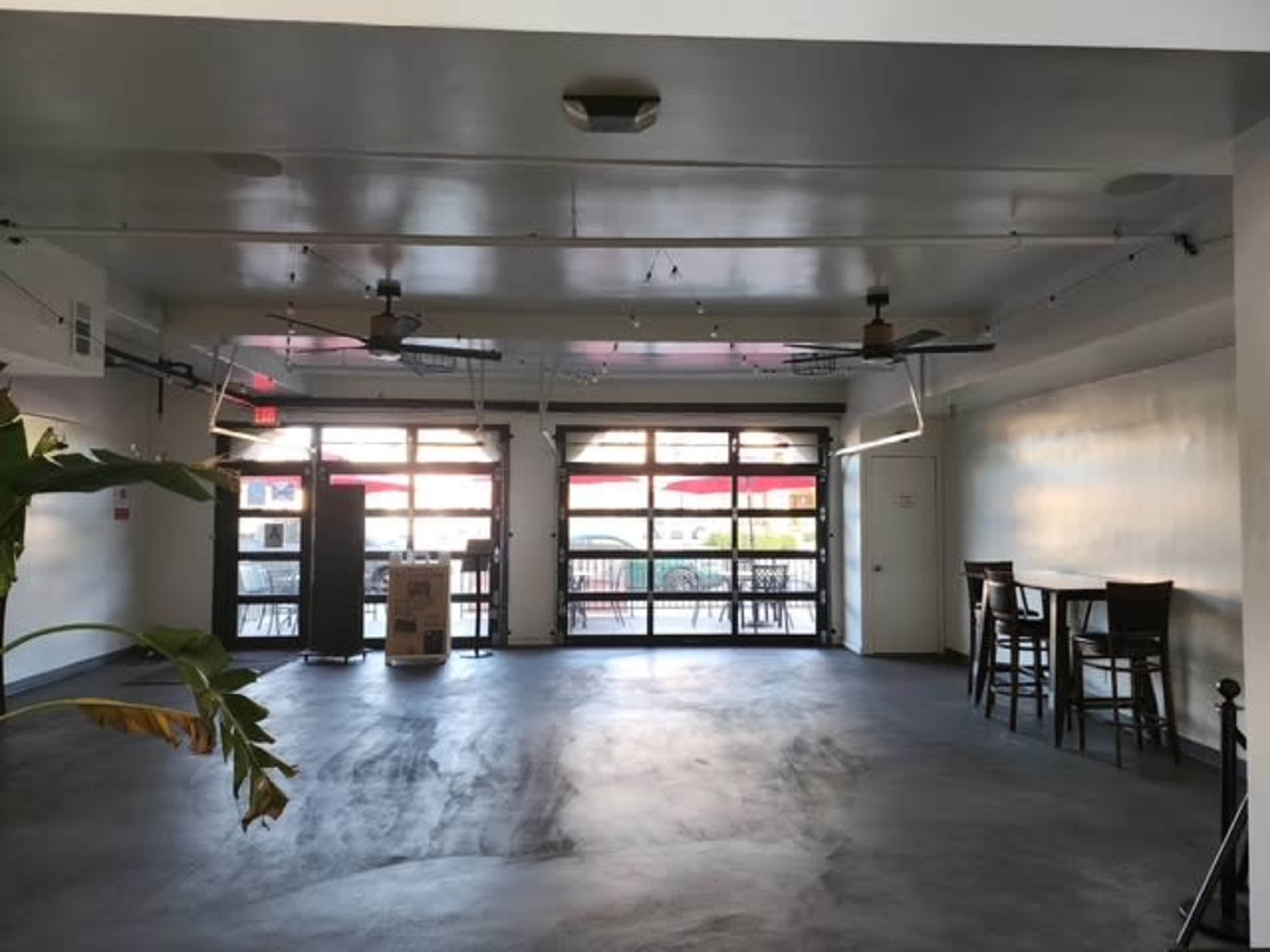 The image shows an empty, spacious interior of a room with large windows, exposed ceiling fans, and a few barstools along the wall.