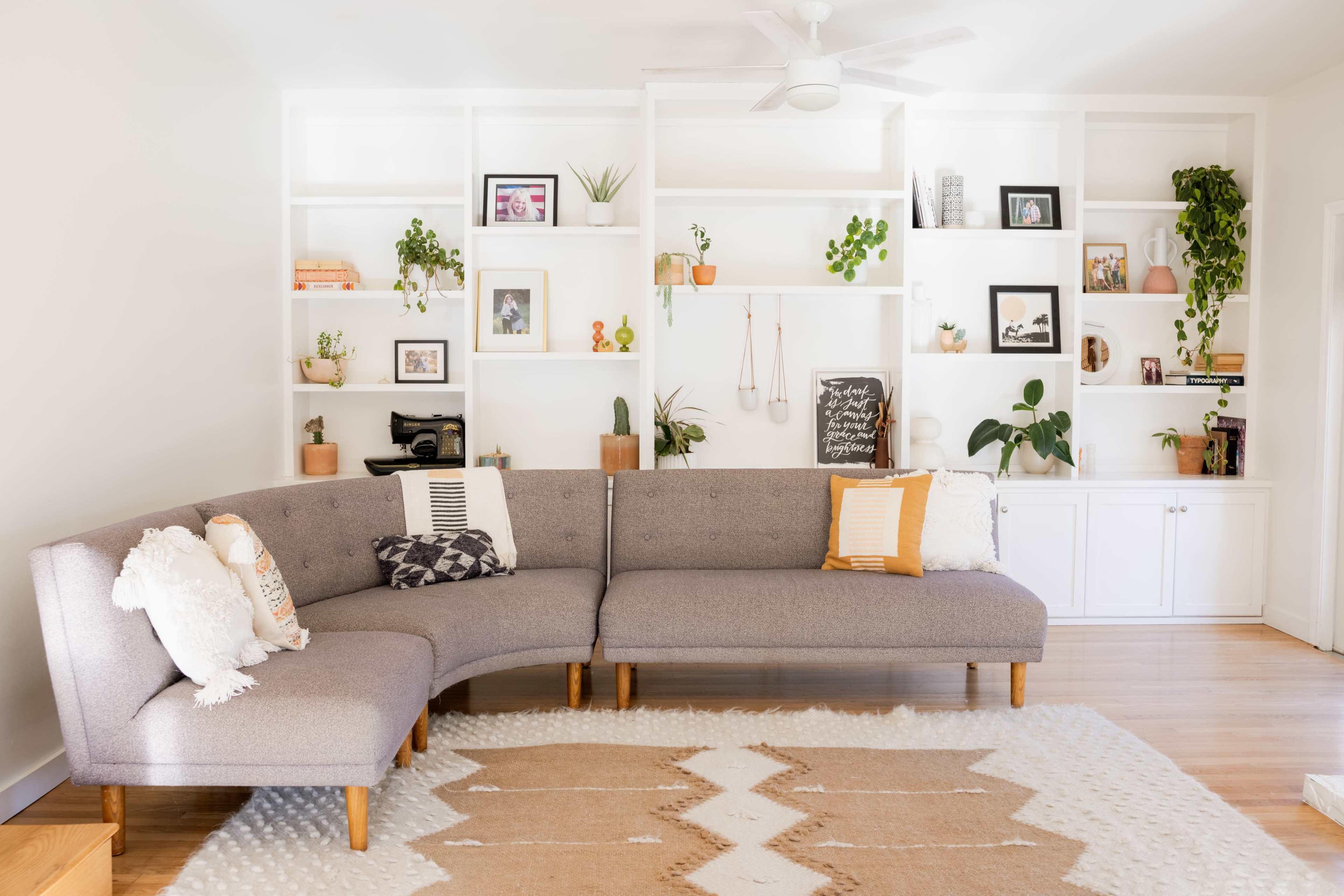 A grey sectional couch with decorative pillows is placed in a well-lit room featuring shelves filled with plants and framed photographs.