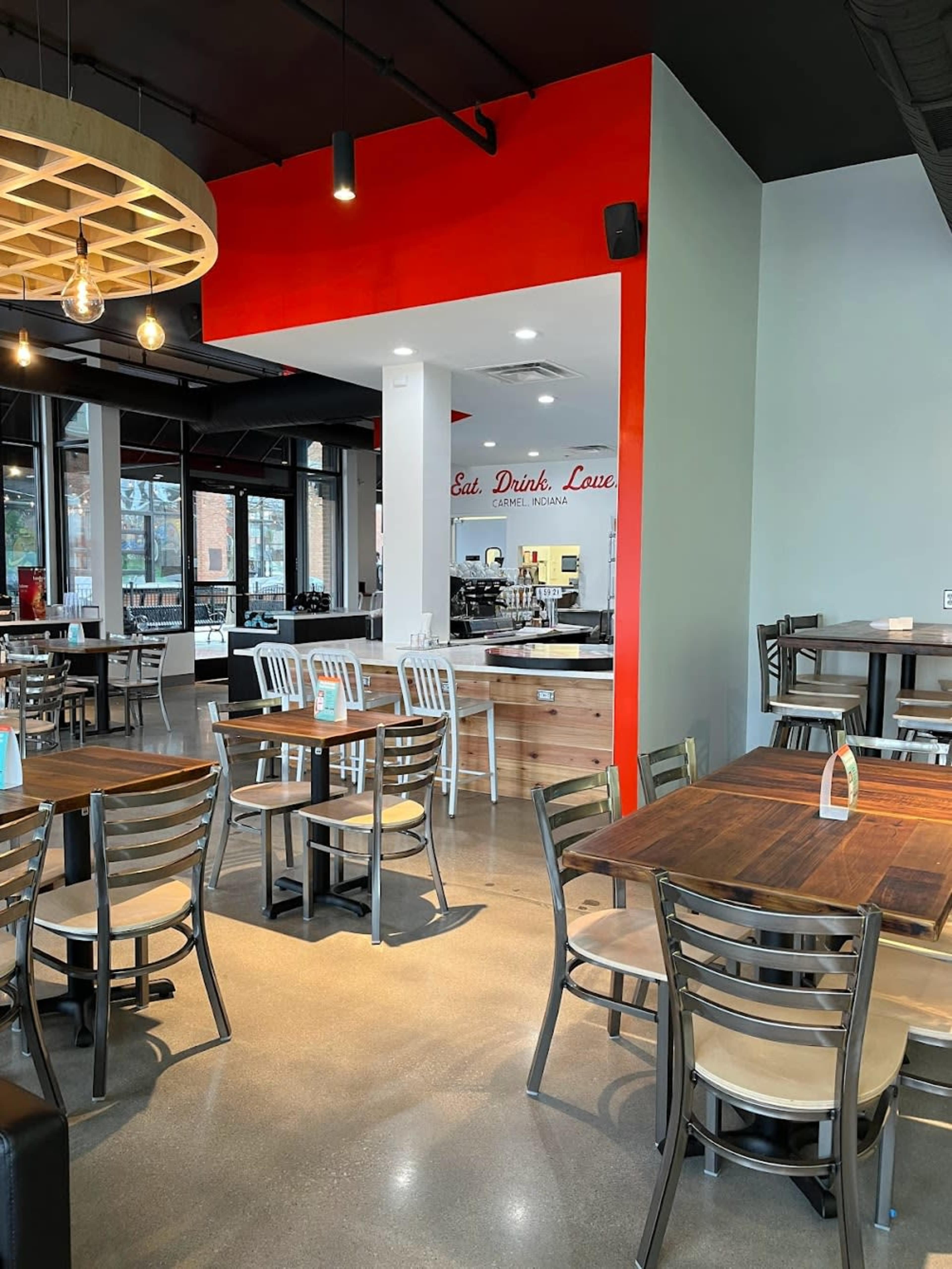 A modern restaurant interior with wooden tables, metal chairs, and a red accent wall.