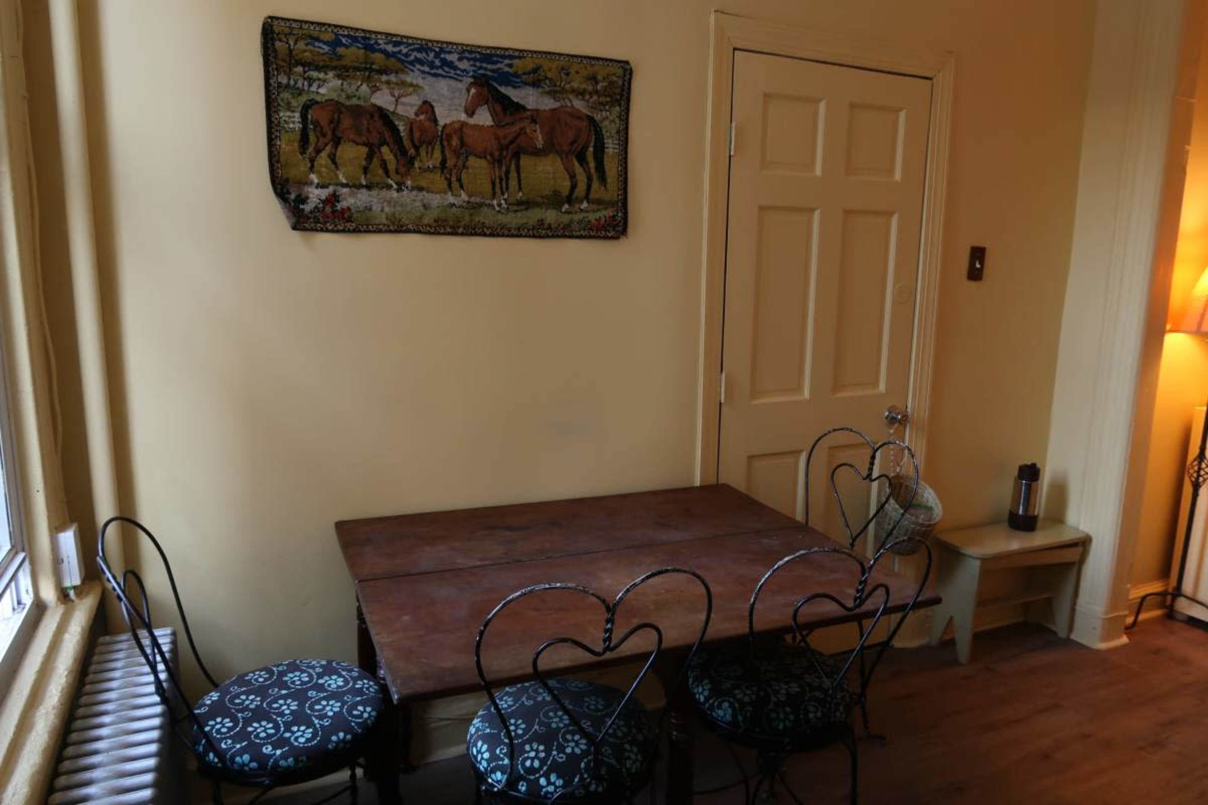 The image shows a small dining area with a wooden table and four metal chairs, adorned with patterned upholstery, next to a wall with a tapestry depicting horses.