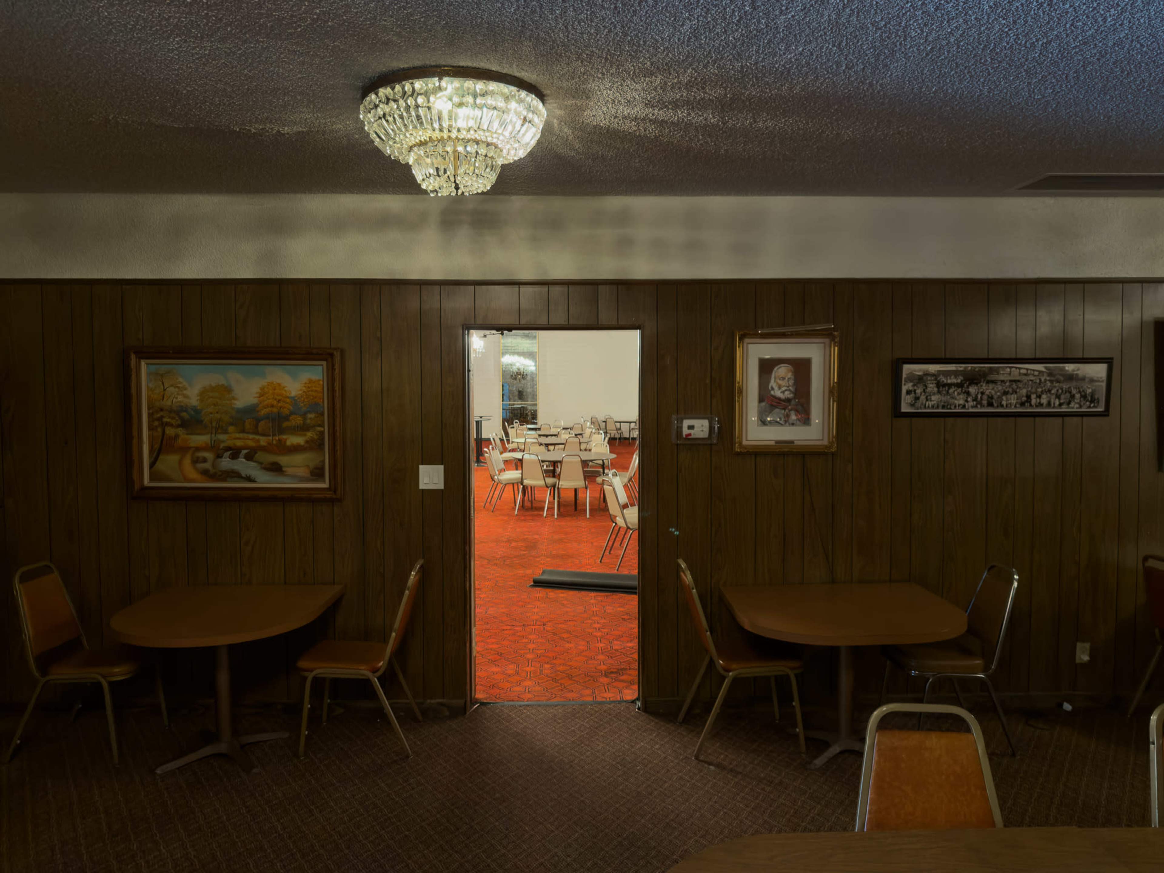A dimly lit room features wooden paneling, empty tables and chairs, and a door leading to a brightly lit space with additional tables and chairs.