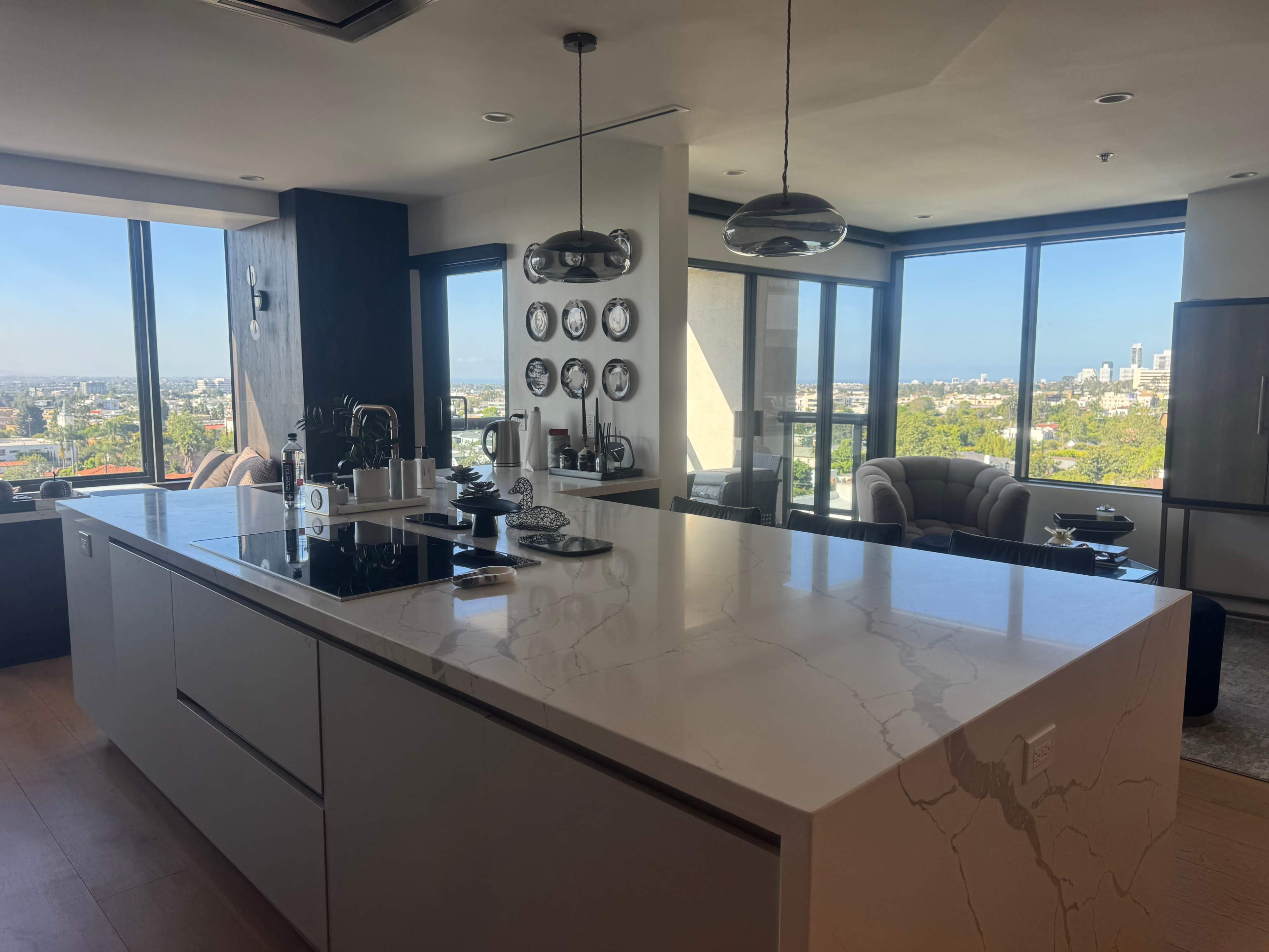 The image shows a modern kitchen with a large island, overlooking a cityscape through expansive windows.