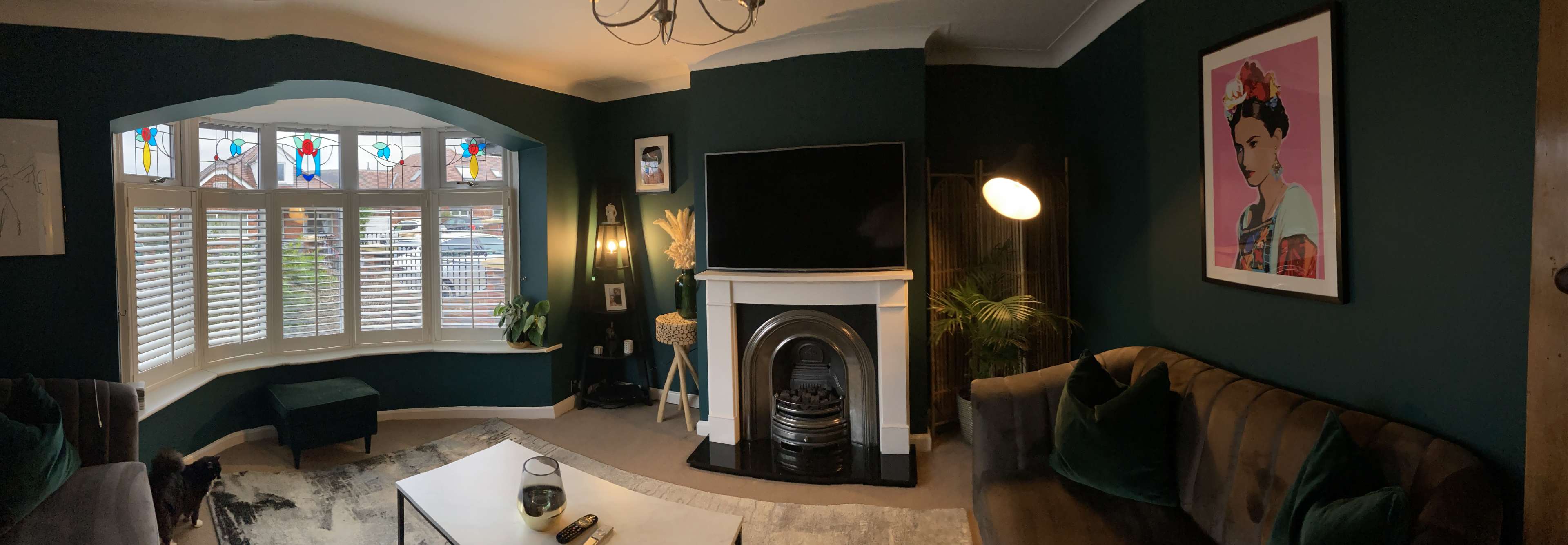 A stylish living room features a dark green accent wall, a large flat-screen TV above a white fireplace, and a bay window with colorful stained glass.