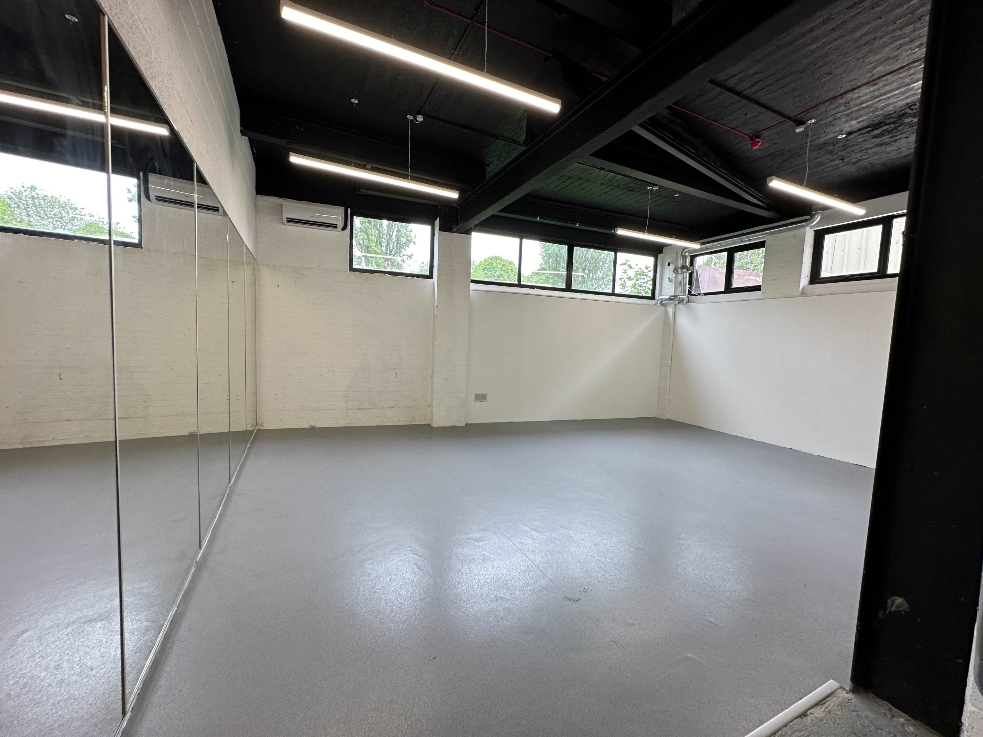 A spacious, bare room with gray flooring and large mirrors on one wall, illuminated by fluorescent lighting.