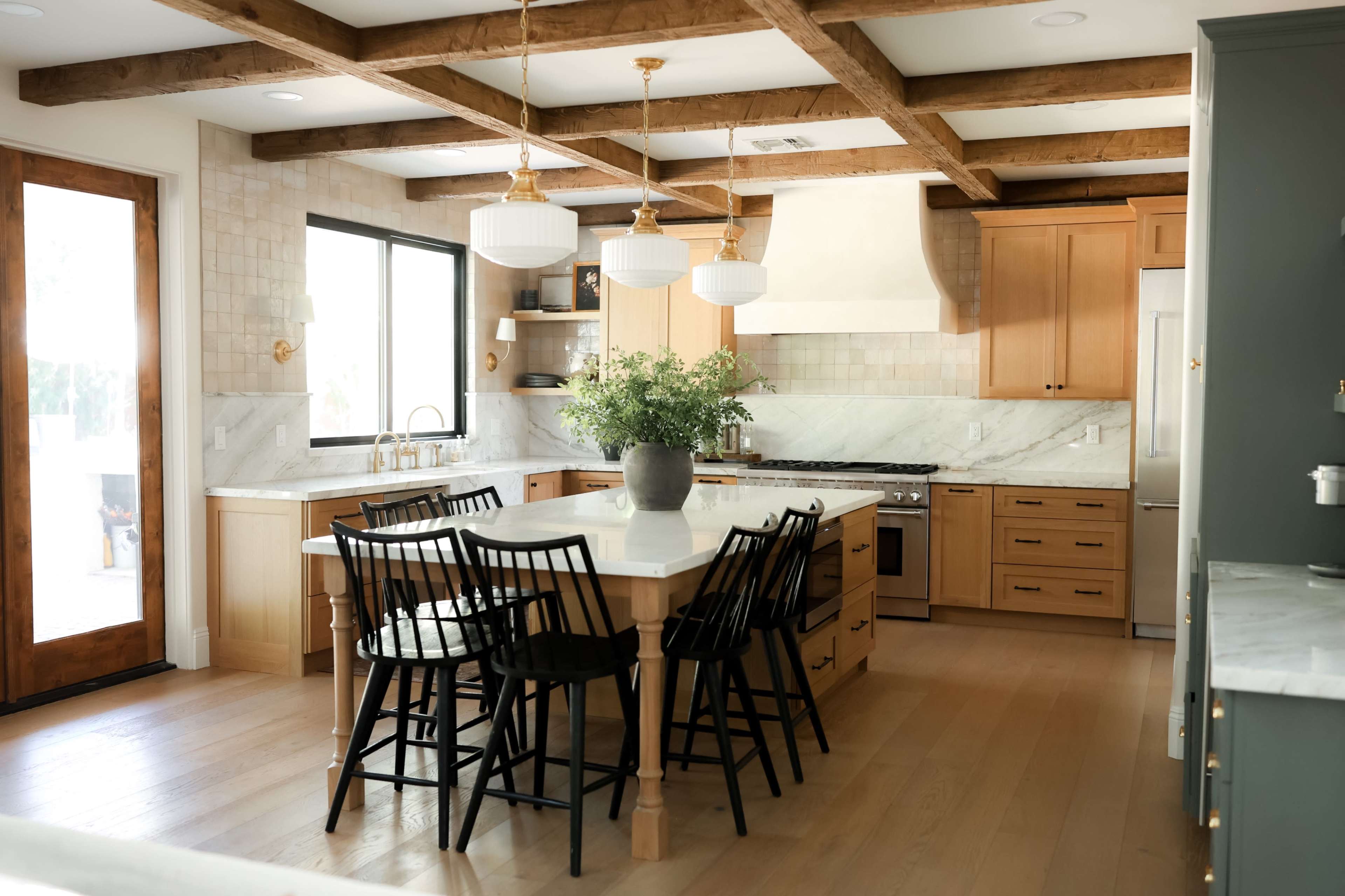 The kitchen features wooden beams on the ceiling, a large island with seating, and light-colored cabinetry paired with marble countertops.