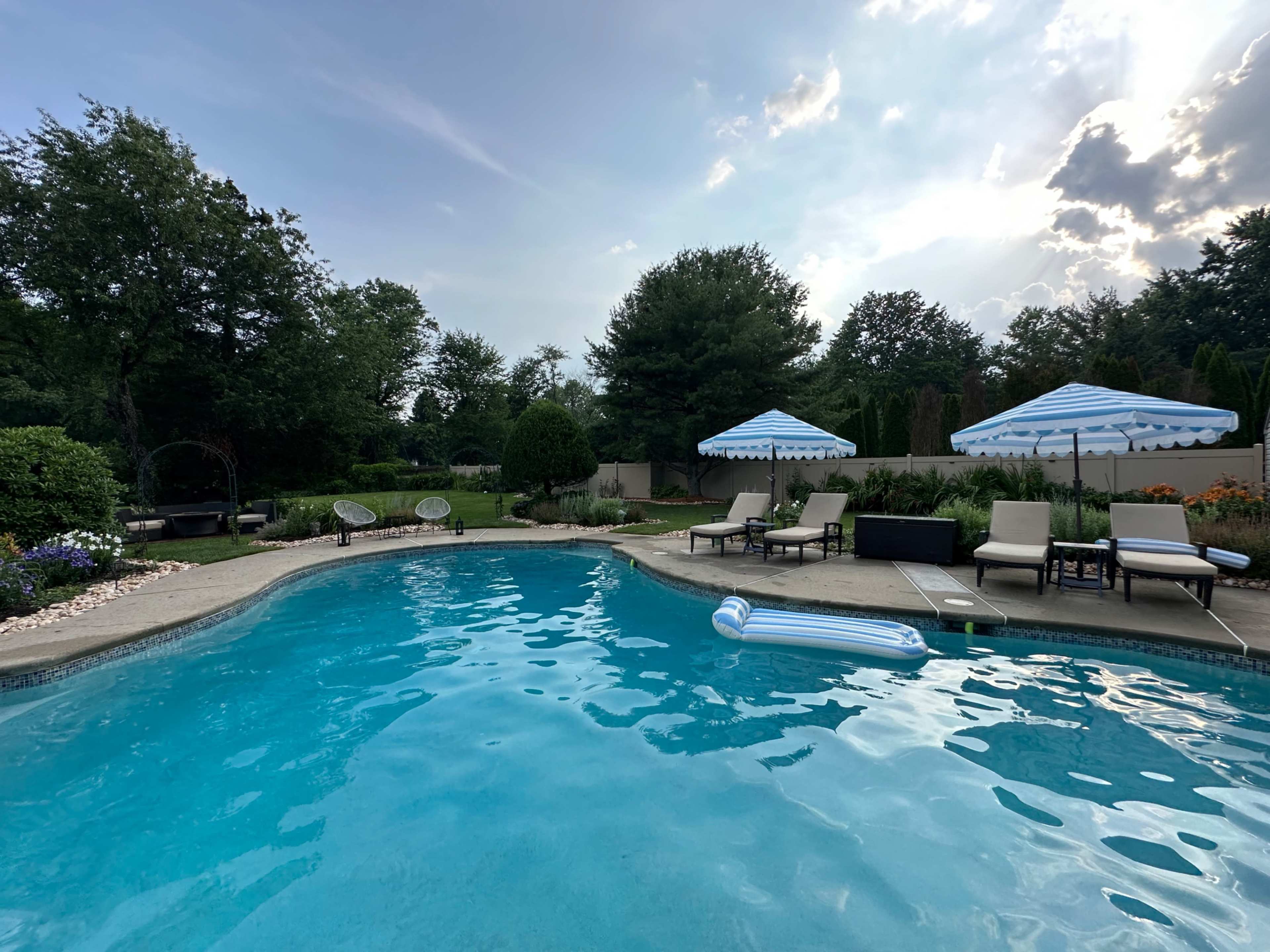 The image depicts a serene backyard pool area with lounge chairs, umbrellas, and surrounding greenery.