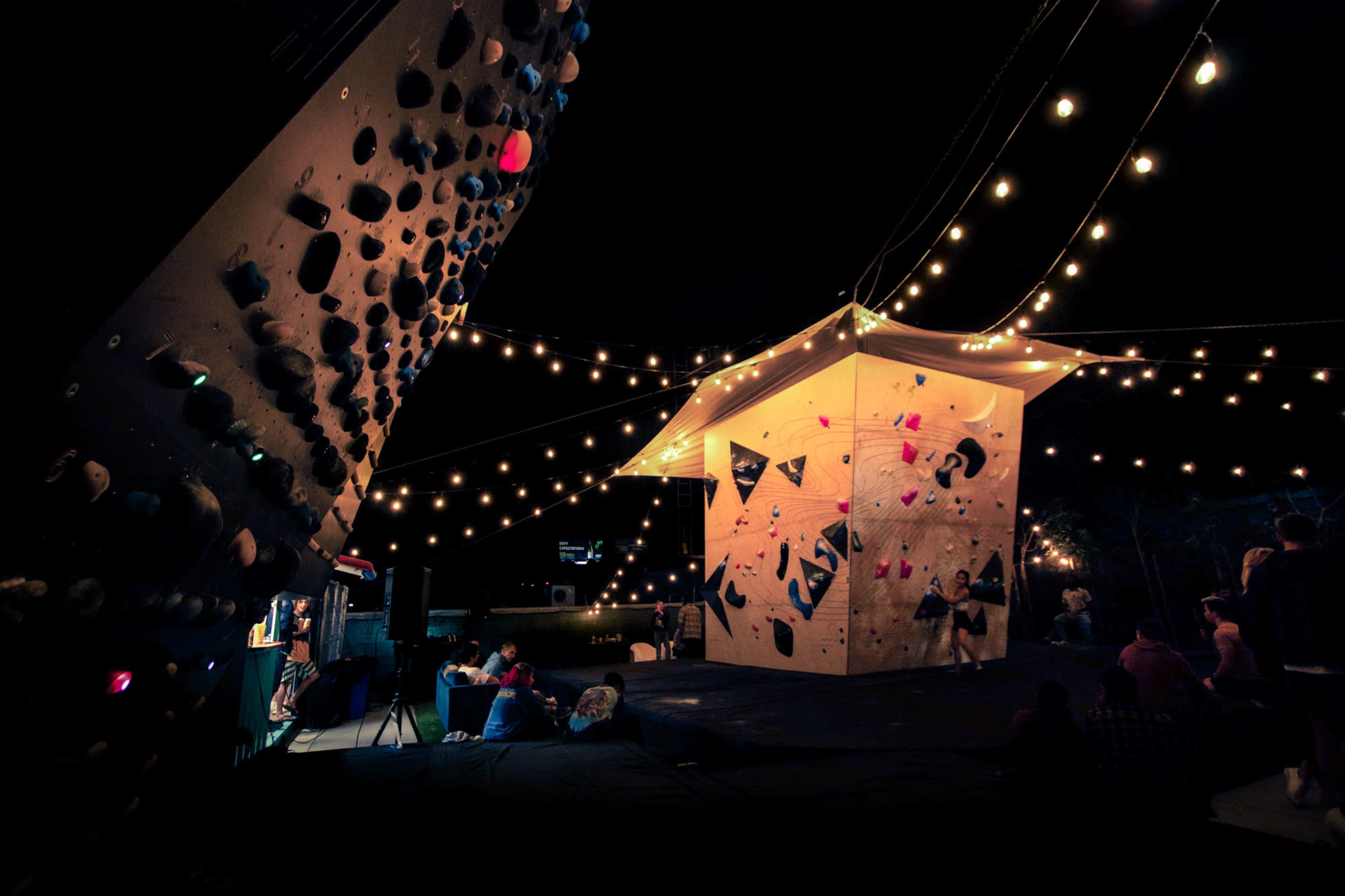 A climbing structure lit by string lights is set up outdoors, with people gathered nearby.