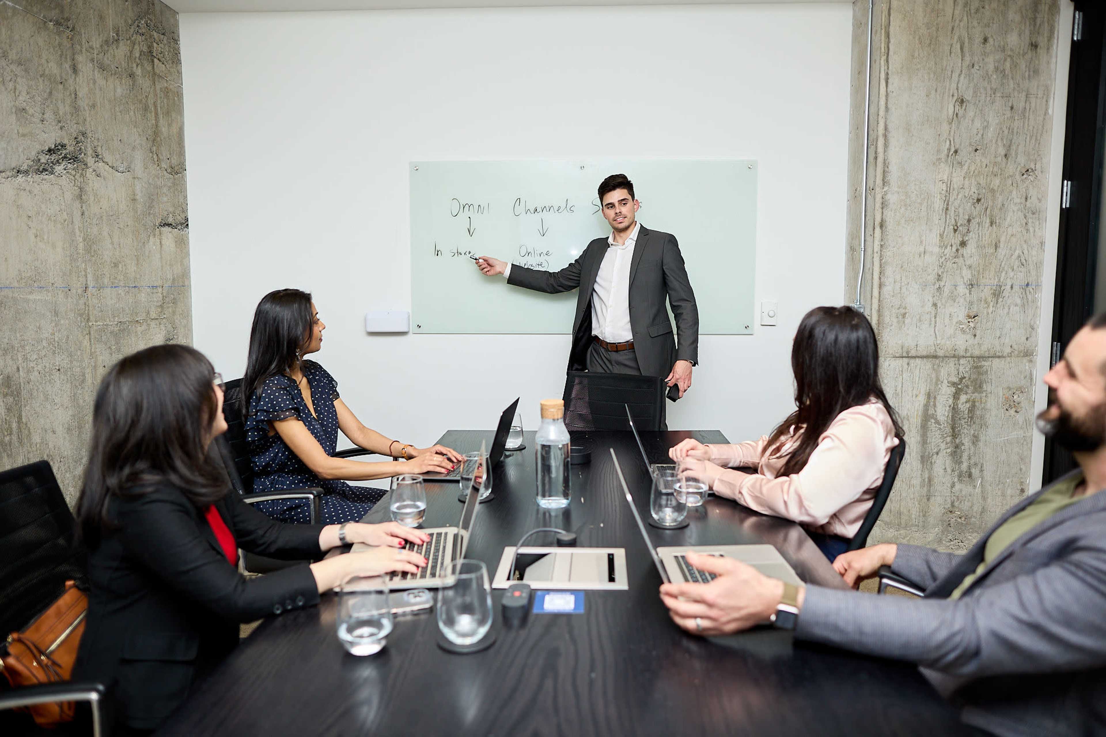 A man in a suit presents information on a whiteboard to a group of colleagues seated at a conference table with laptops and water glasses.