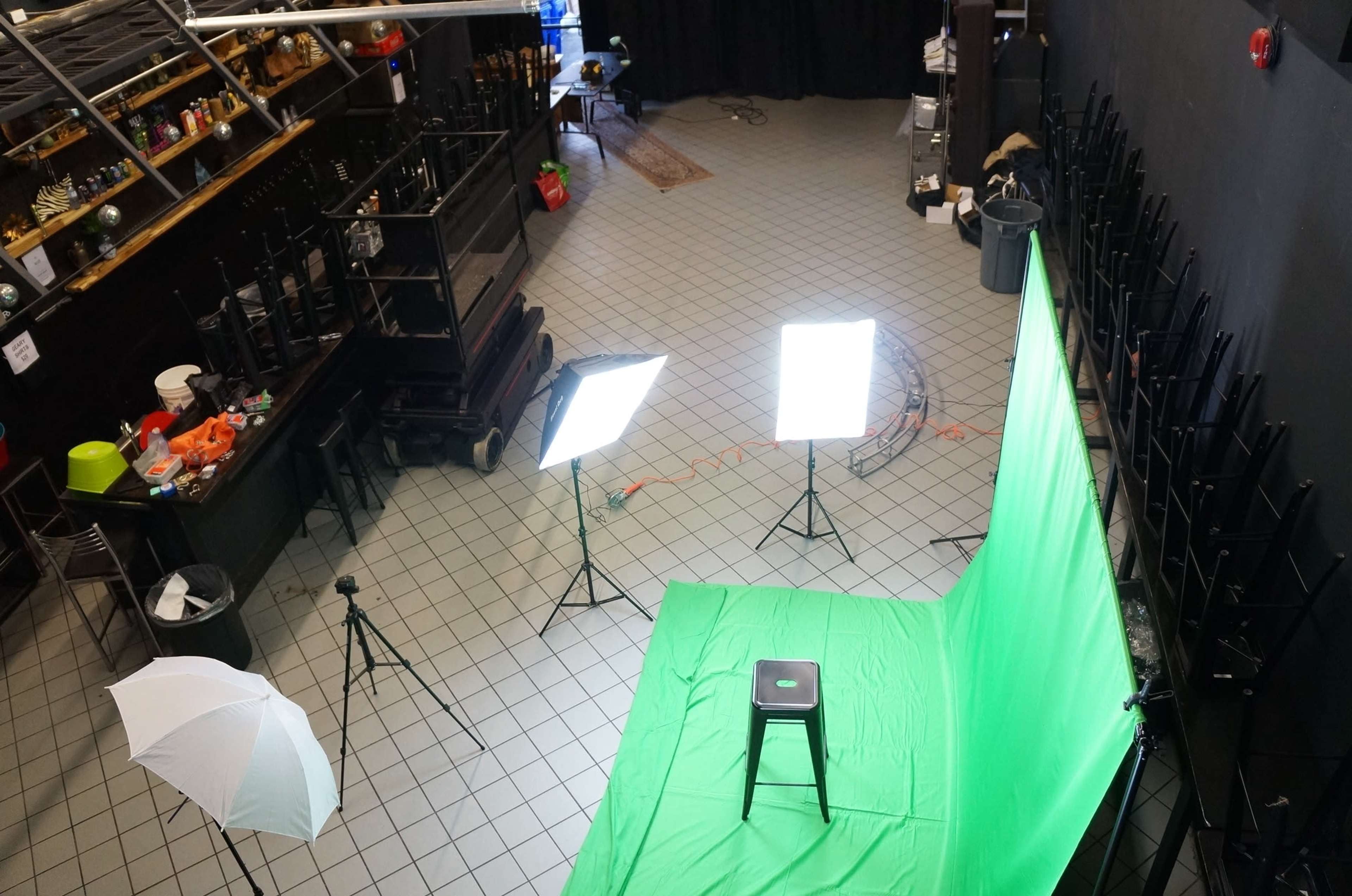 A photography studio setup with a green screen, lighting equipment, and a stool positioned in front of it, all in a tiled space with stacked chairs and various props.