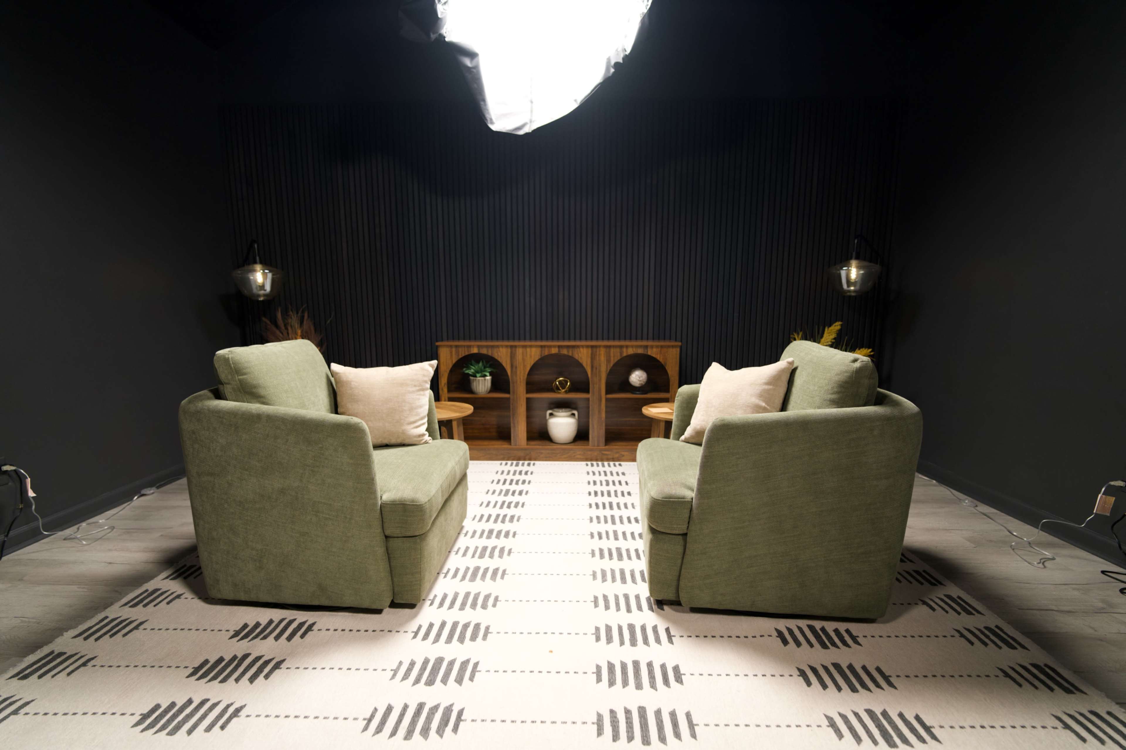 The image shows two green sofas facing each other on a patterned rug, with a wooden shelf and decorative items in the background against a dark wall.
