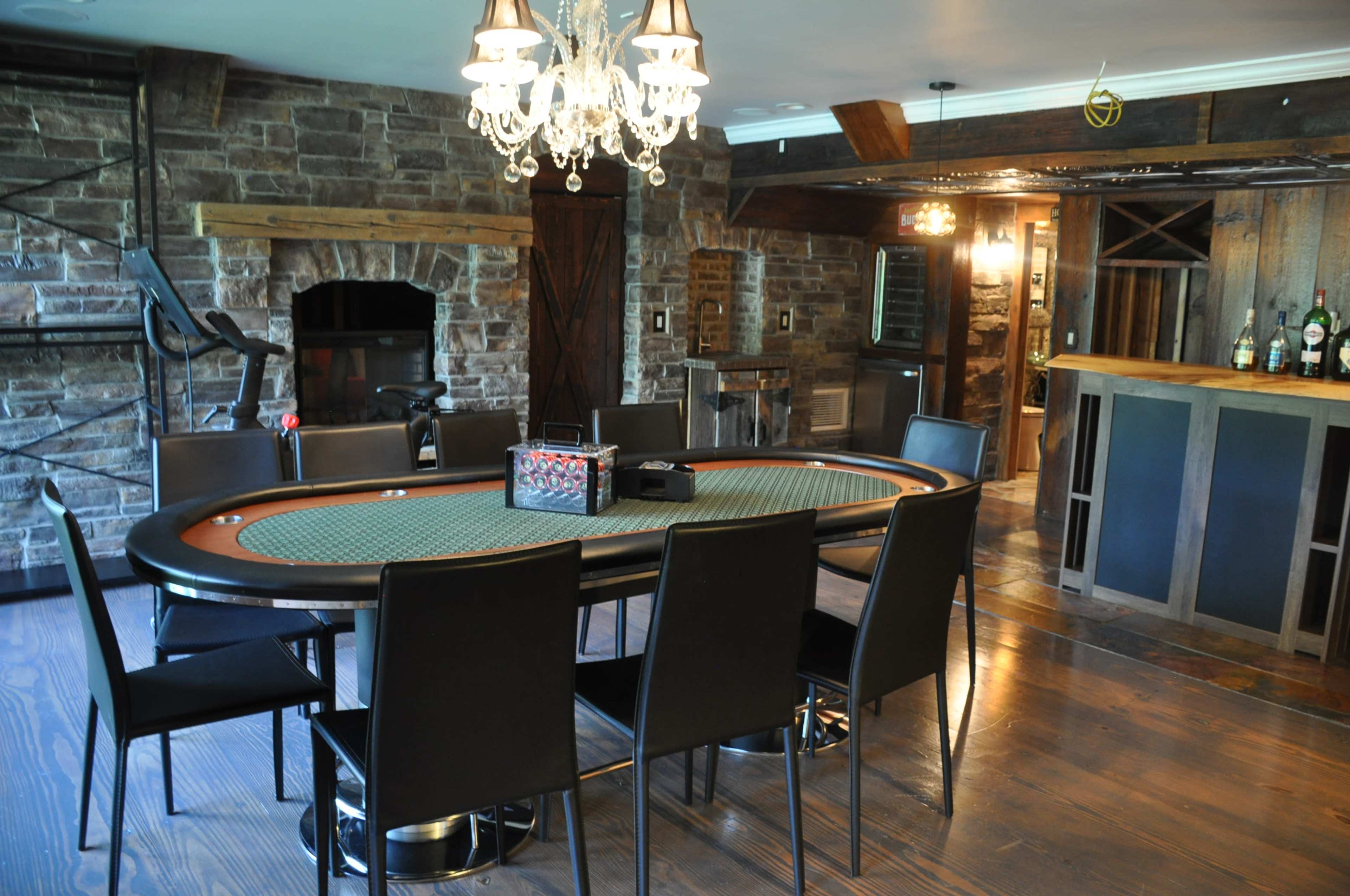 The image shows a poker table surrounded by black chairs in a room with stone walls, a chandelier, and a bar area in the background.