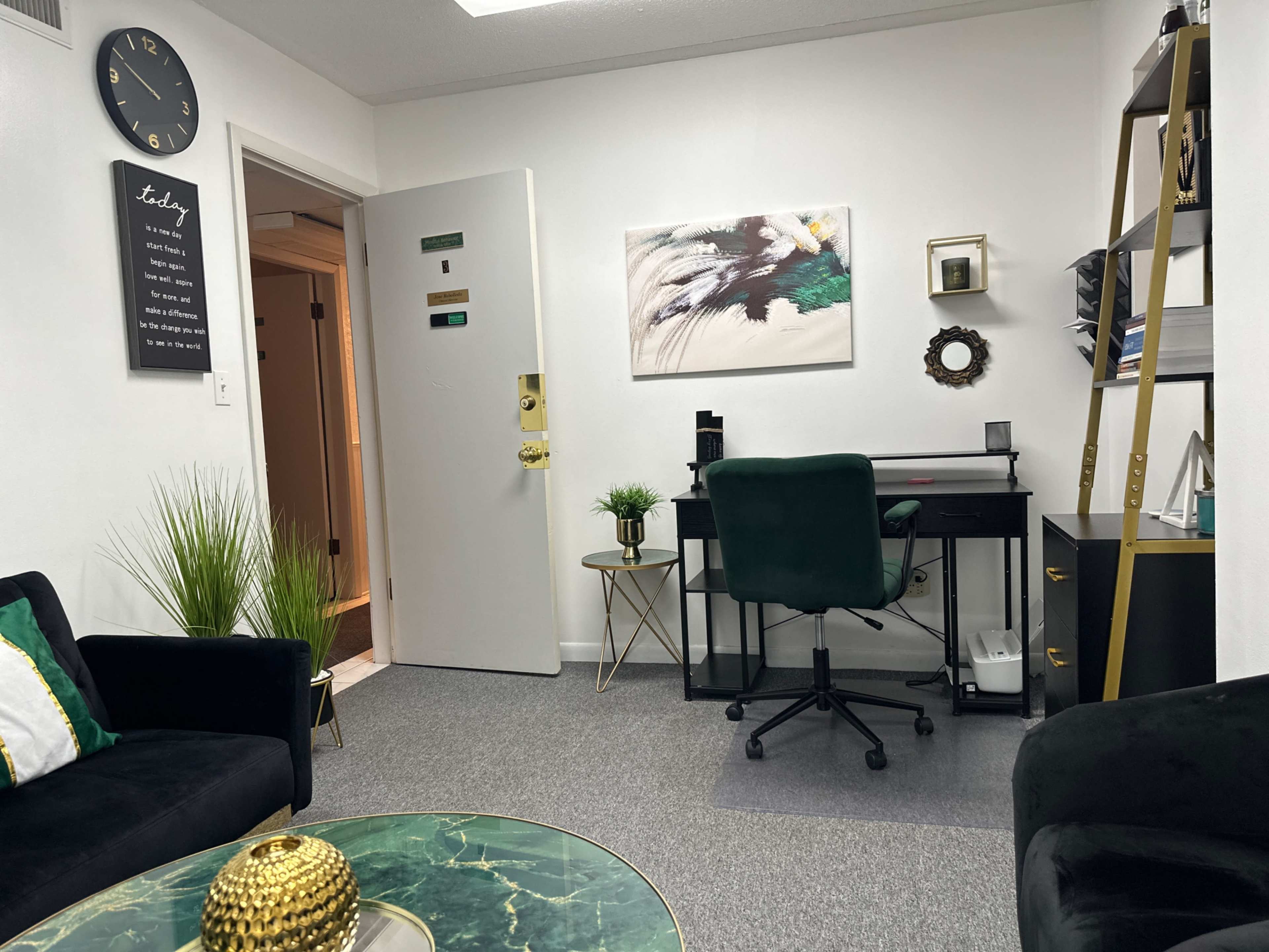 A modern office space featuring a desk, a chair, plants, and a green accent wall with decor, along with a clock and an entrance door.