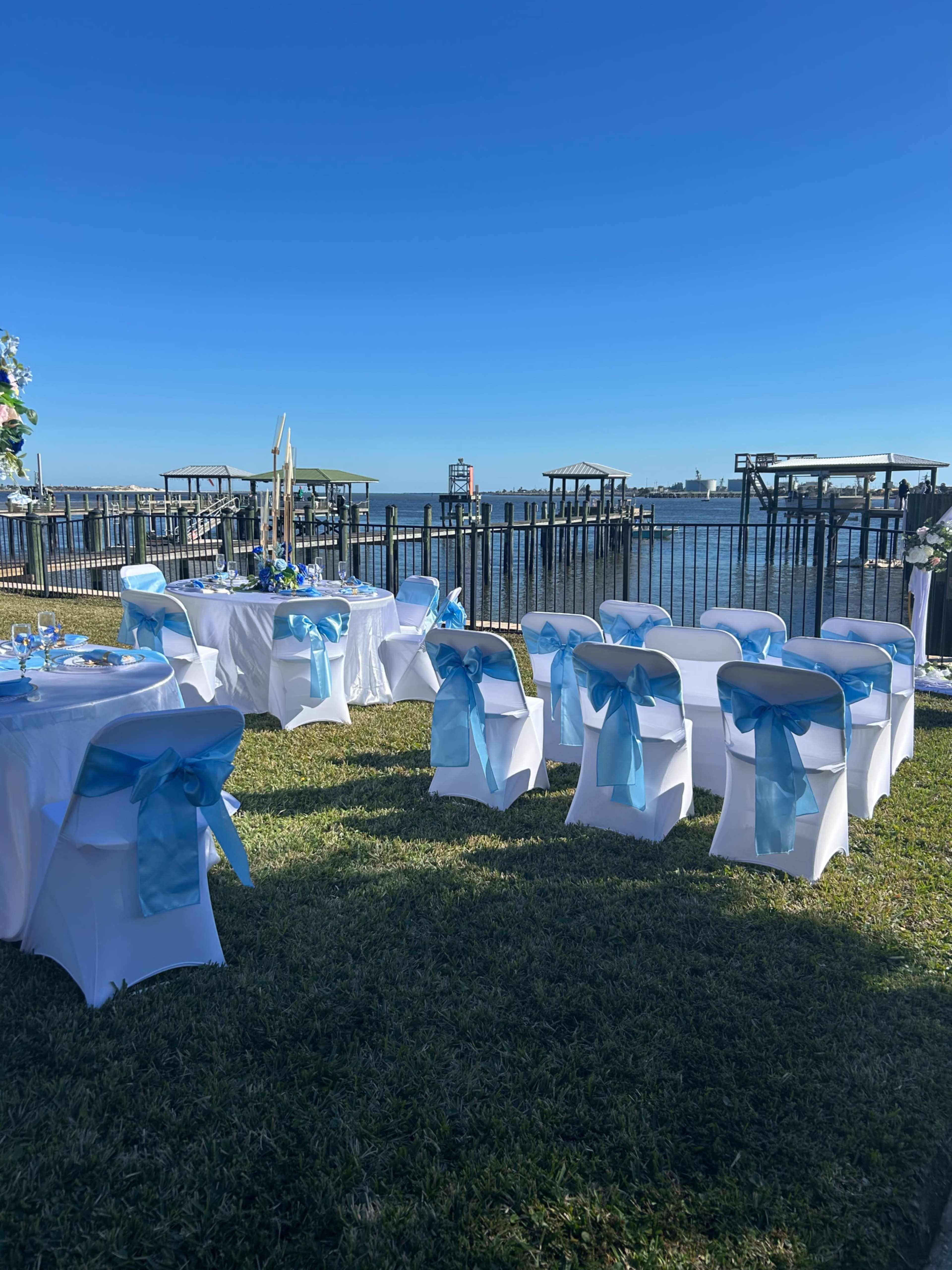 The outdoor venue features neatly arranged tables and chairs adorned with blue sashes, overlooking a body of water and a series of boat docks in the background.