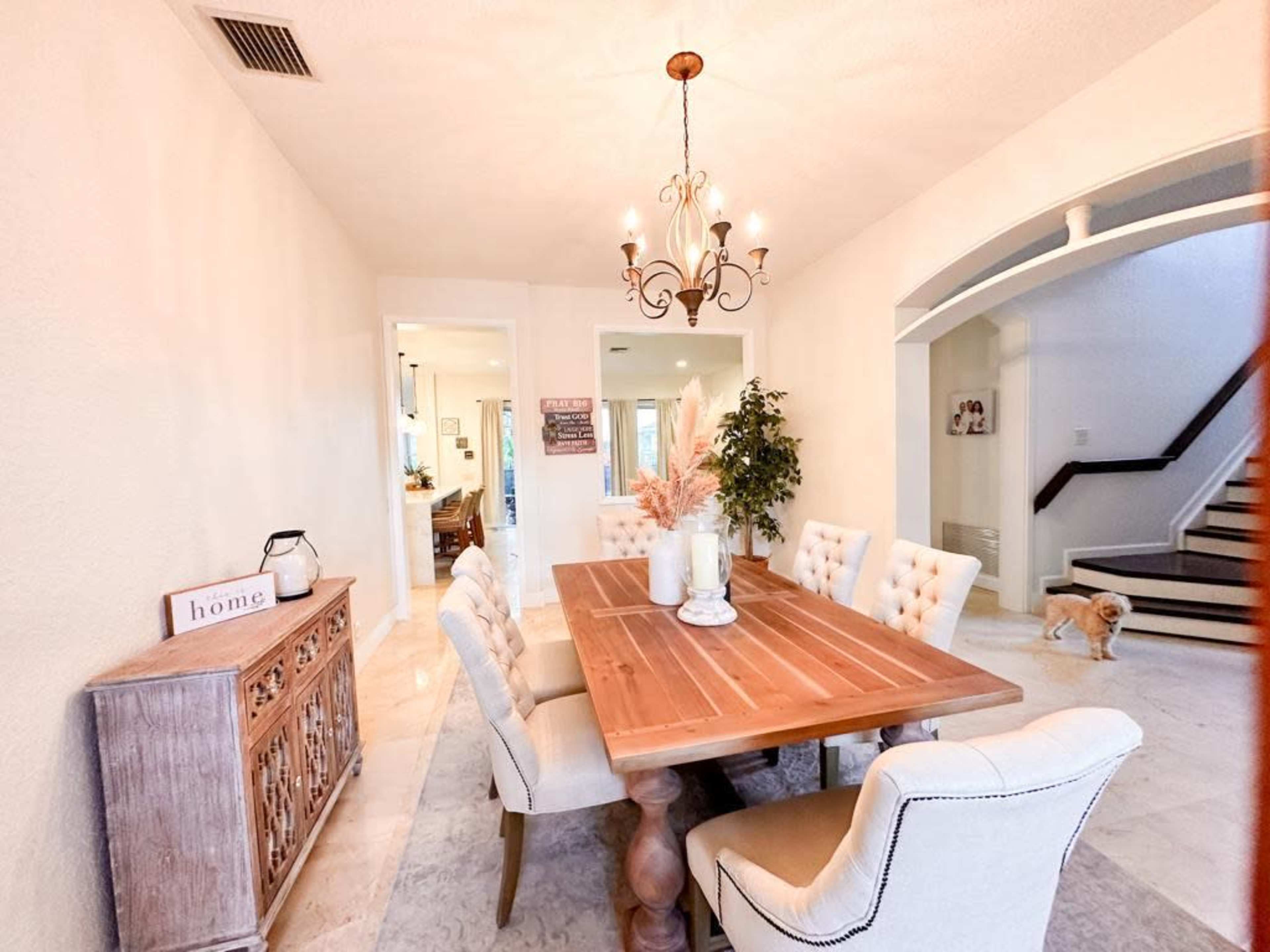 A wooden dining table surrounded by upholstered chairs is set in a well-lit room with a chandelier, leading to a staircase and another living area.