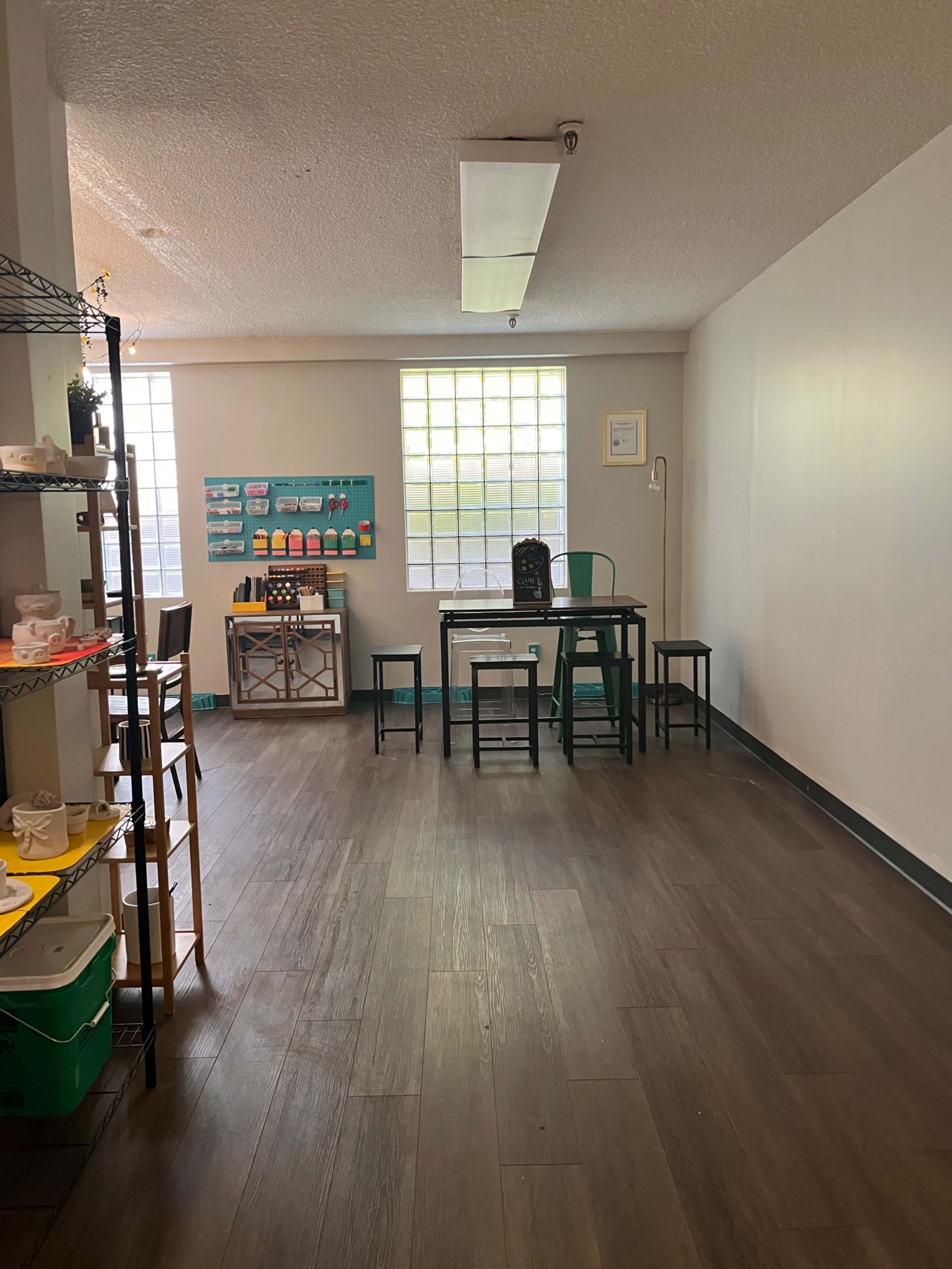 The image shows a bright, spacious room with several tables and chairs, a wall display, and a shelf filled with various items.