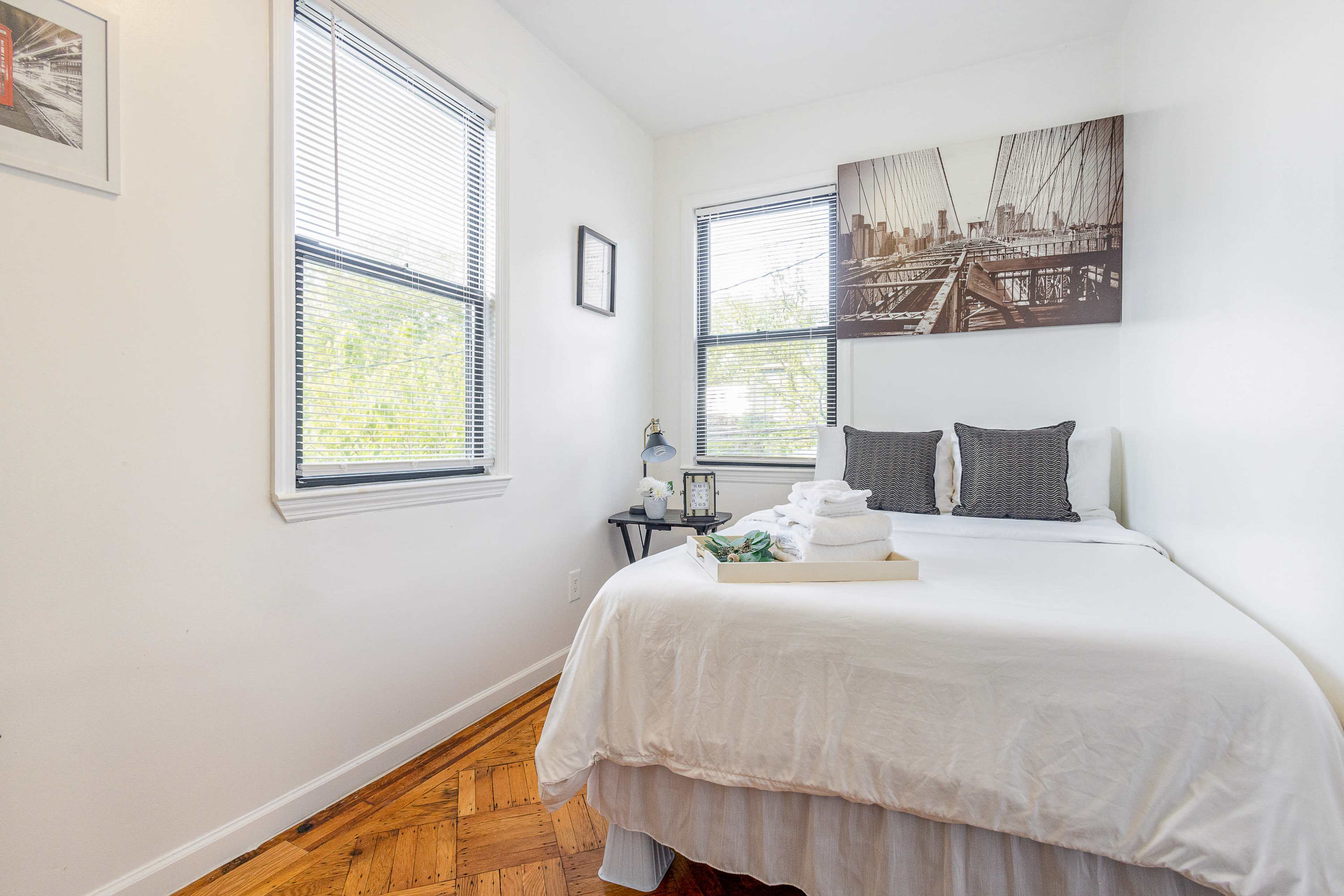 A small bedroom features a bed with white linens, a tray of folded towels, and two windows allowing natural light.