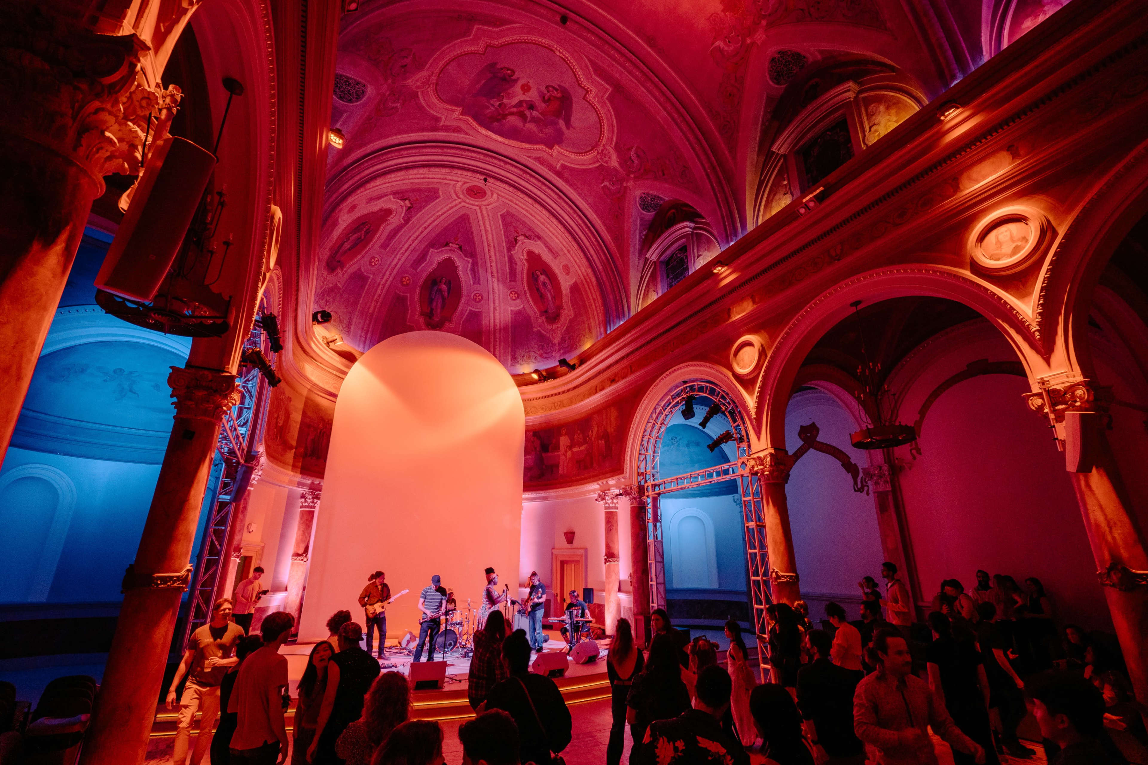 A band performs on stage in a brightly lit, ornate hall filled with an audience.