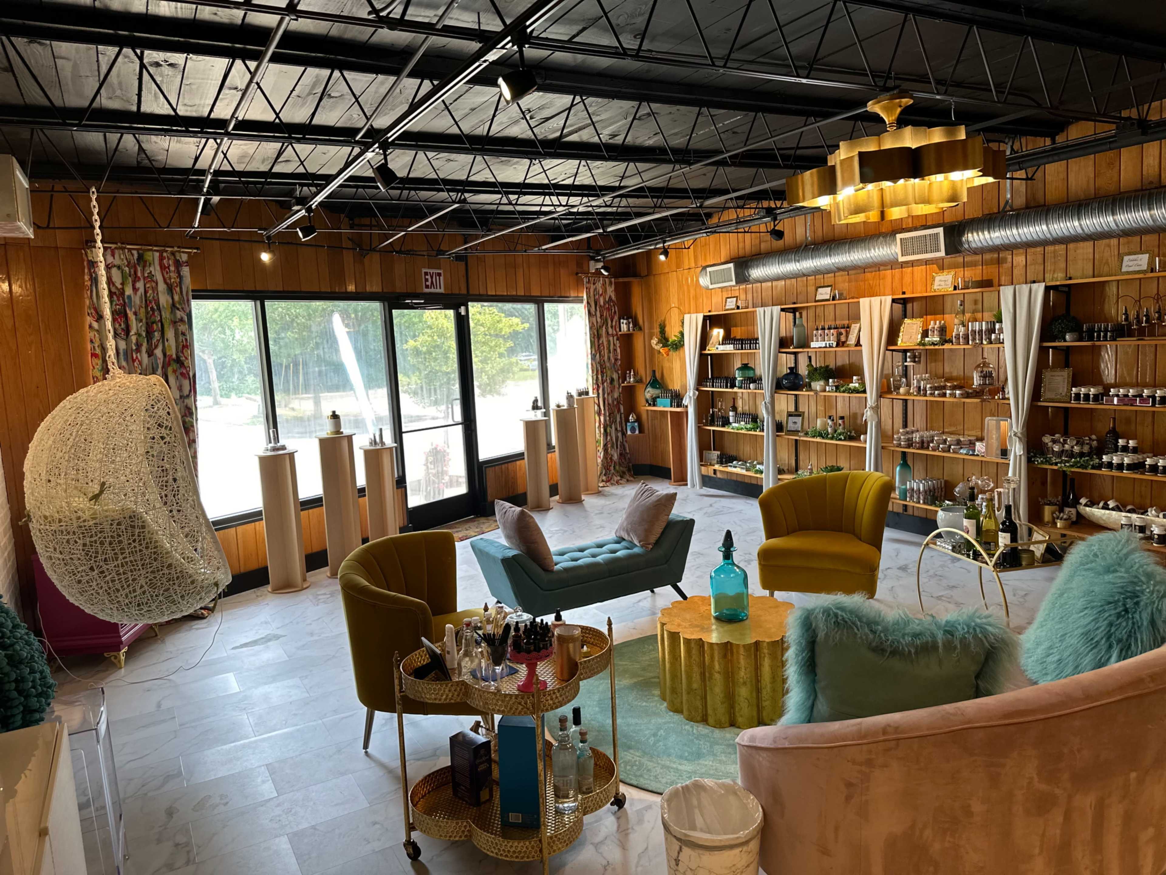 The image shows a well-decorated interior of a shop featuring various displays of products arranged on wooden shelves, with seating areas that include green and pink chairs and a round coffee table.