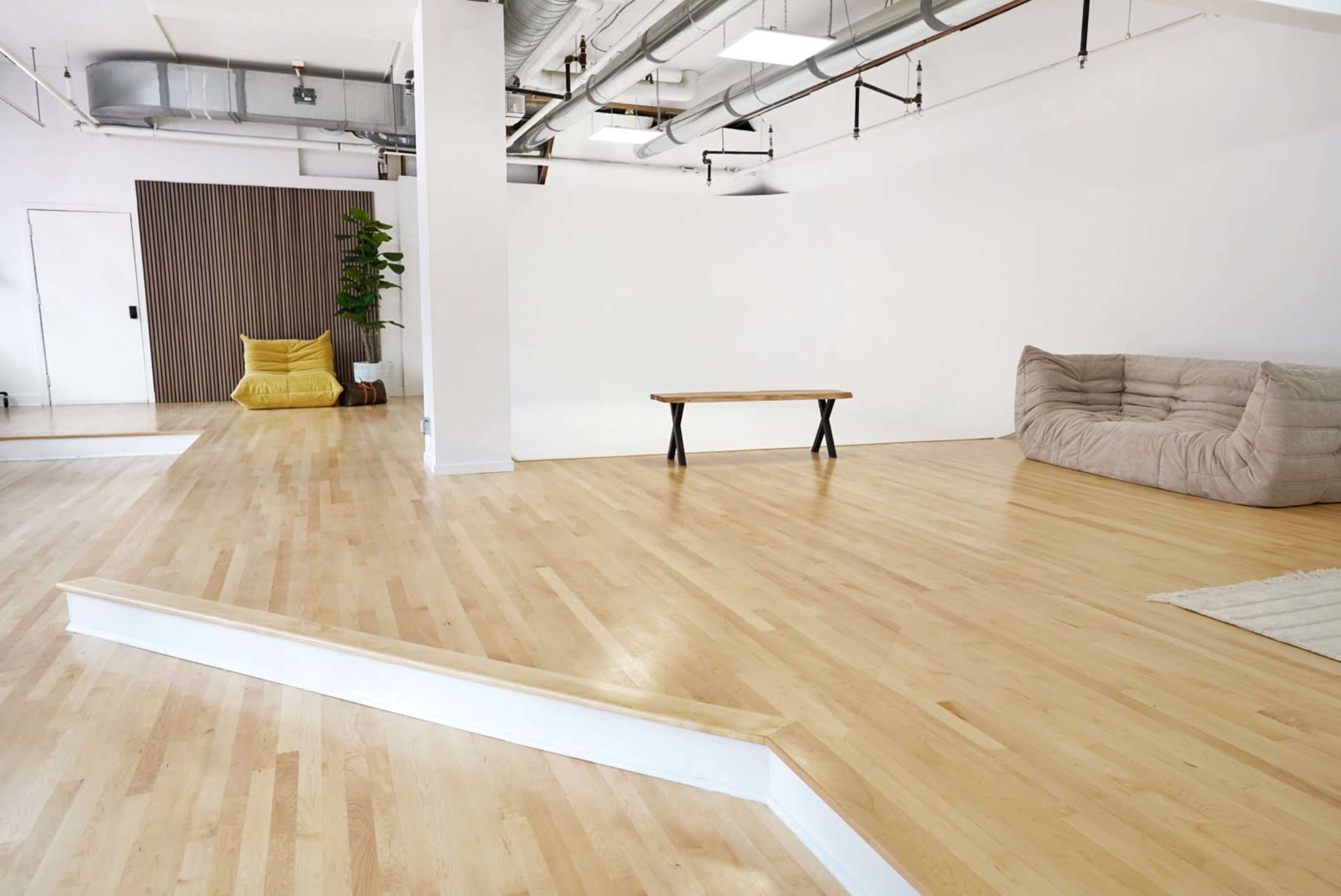 An empty room features wooden flooring, a large sofa, a coffee table, and a small plant against a wall.
