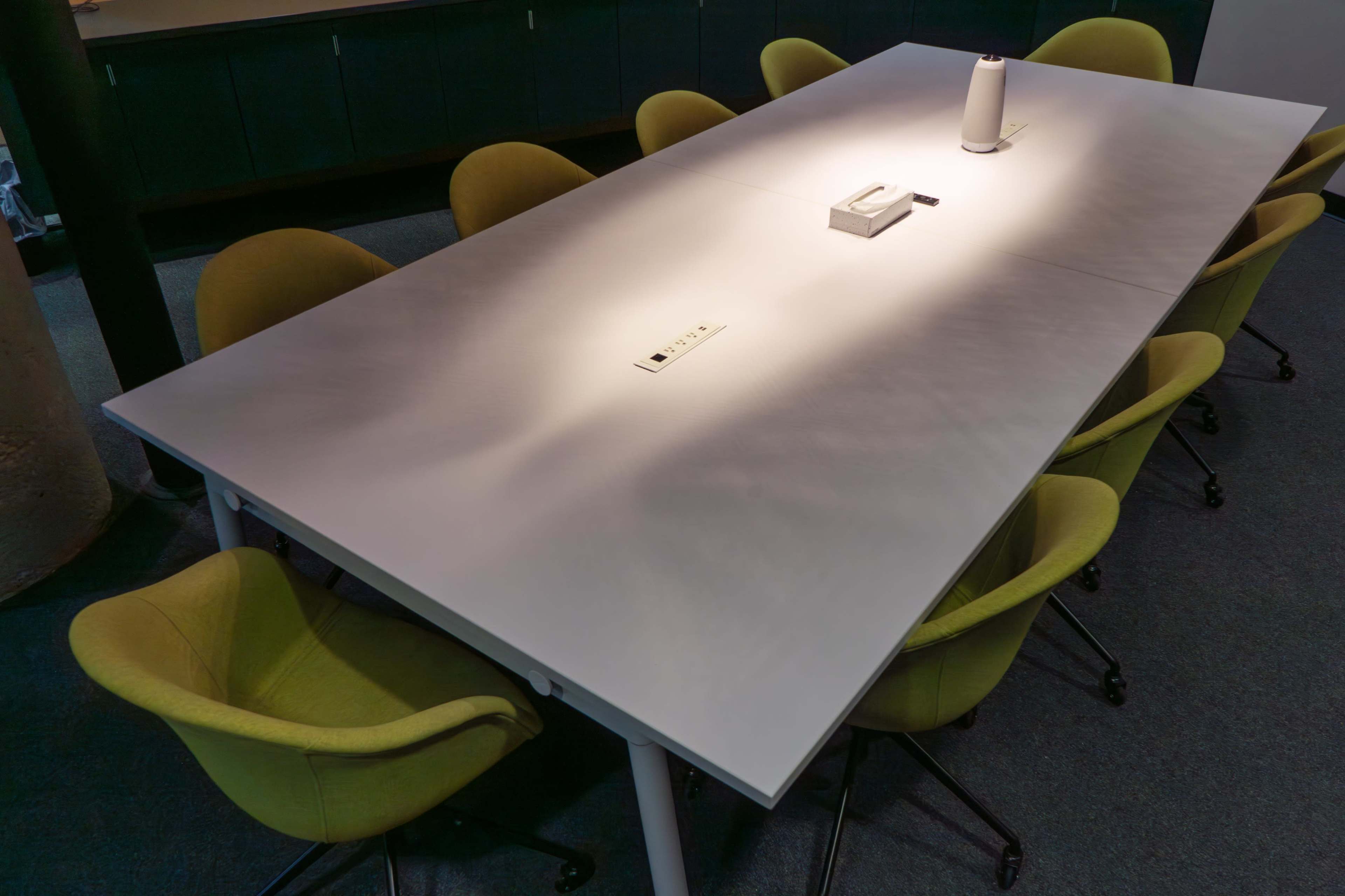 A long, rectangular conference table with a single central light fixture is surrounded by several green chairs in a modern office setting.