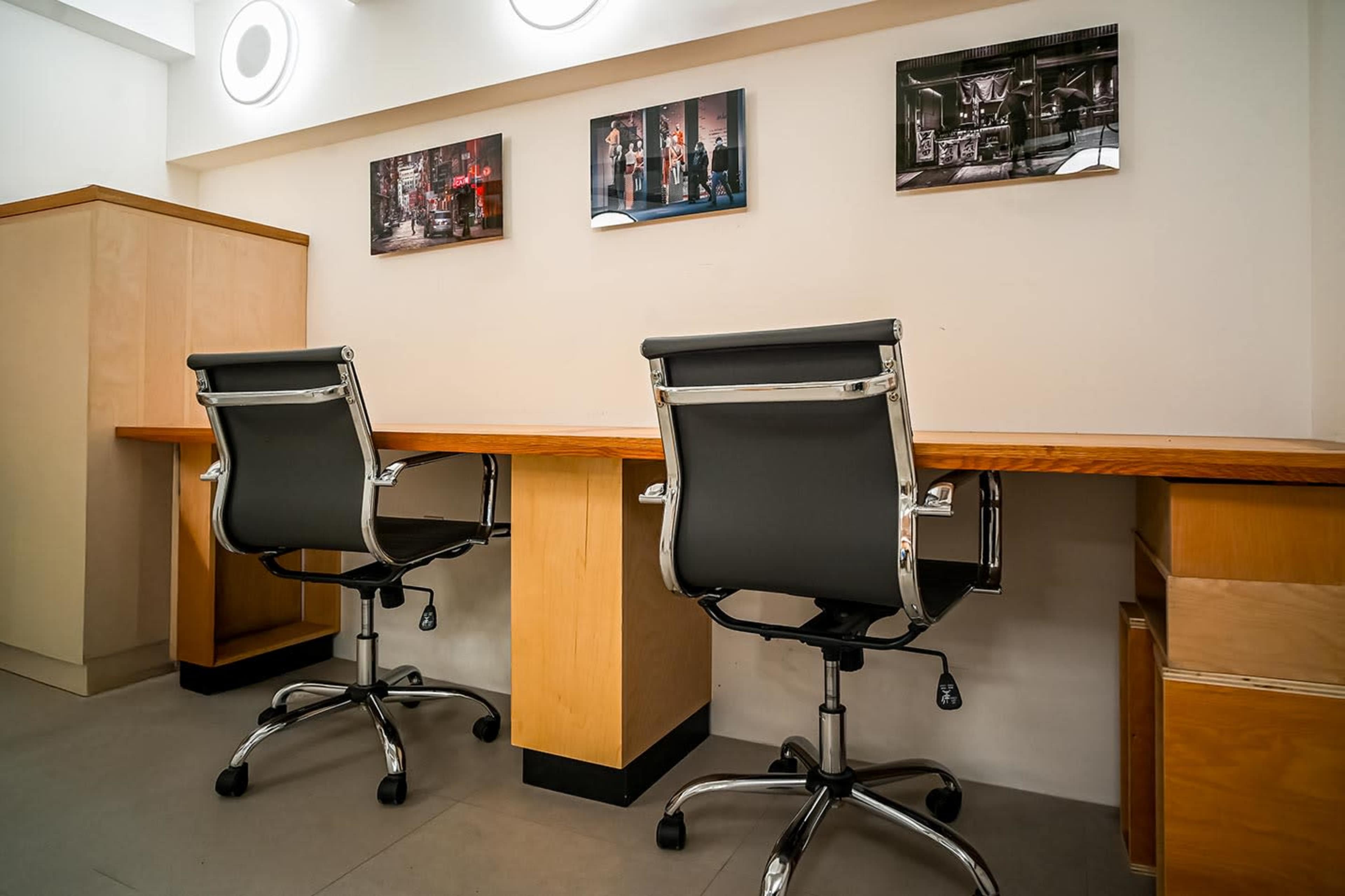 The image shows a minimalist office space with two empty black office chairs positioned at a wooden desk, accompanied by framed photographs on the wall above.