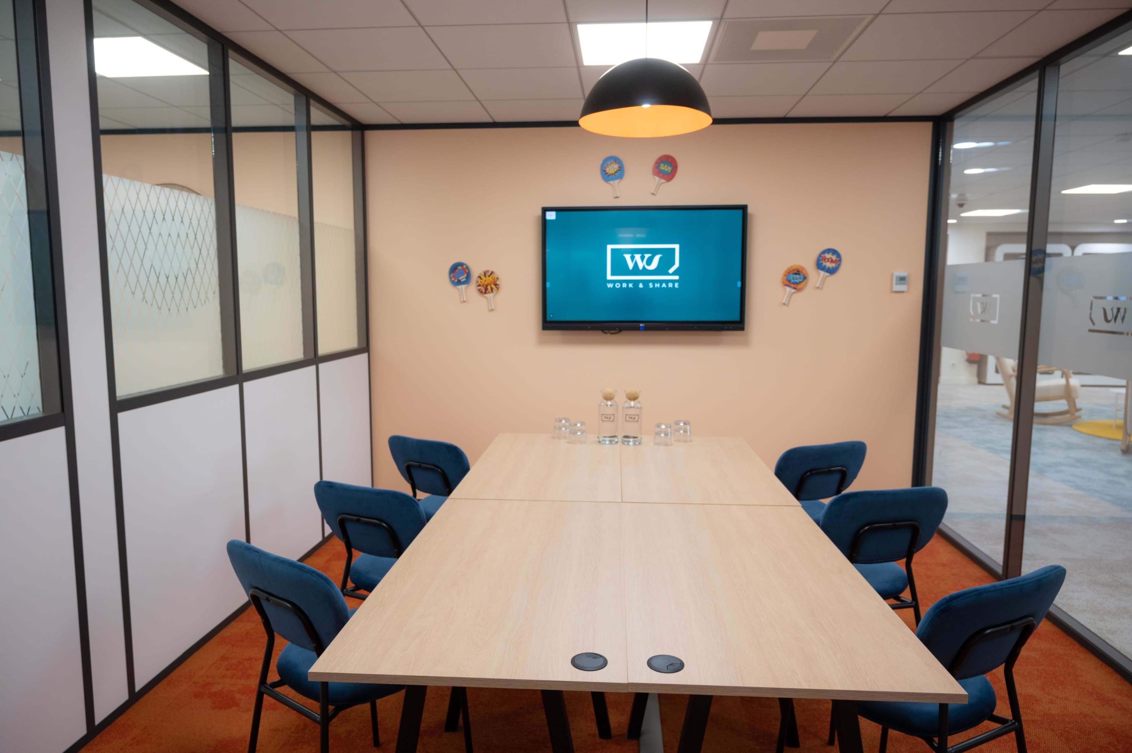 A modern conference room with a large wooden table, six blue chairs, and a wall-mounted television displaying a logo.