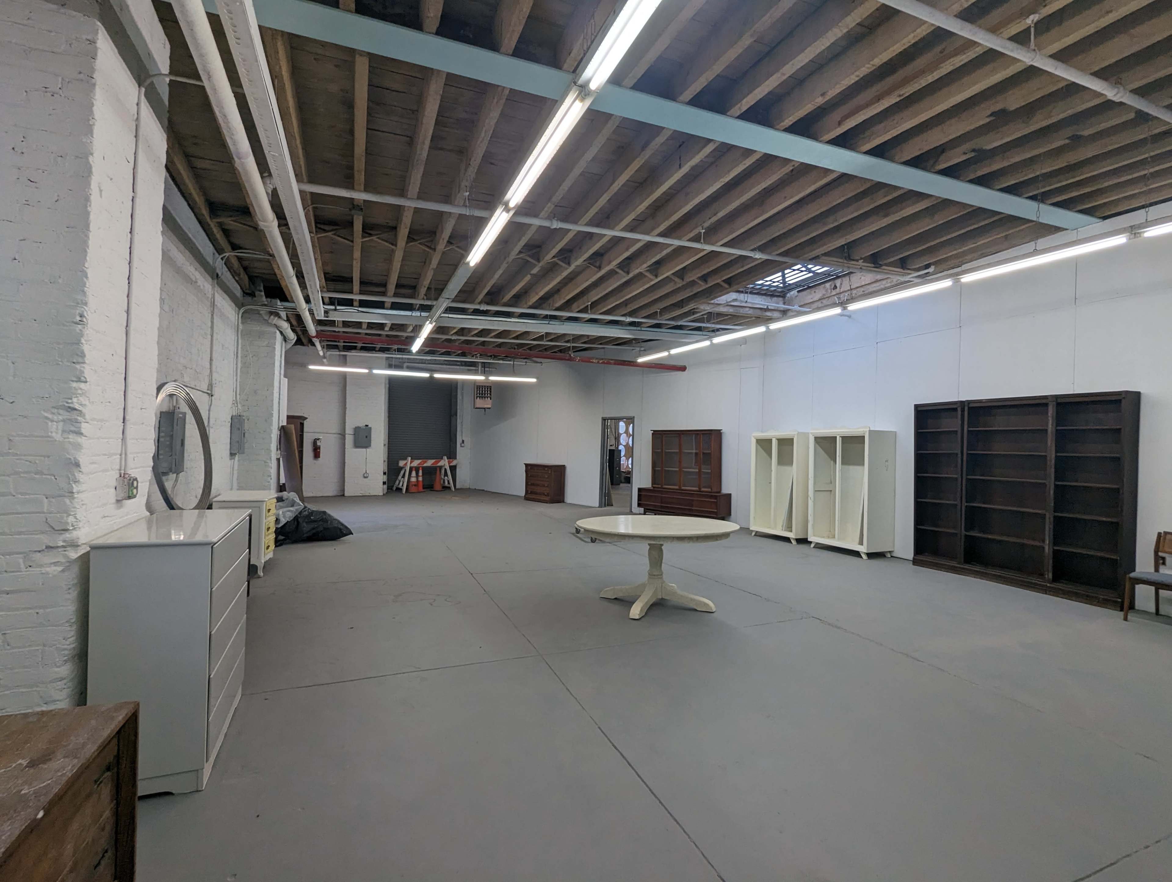 The image shows a spacious empty room with hardwood beams on the ceiling, featuring several pieces of furniture including a round table, white shelving units, and dark wooden cabinets.