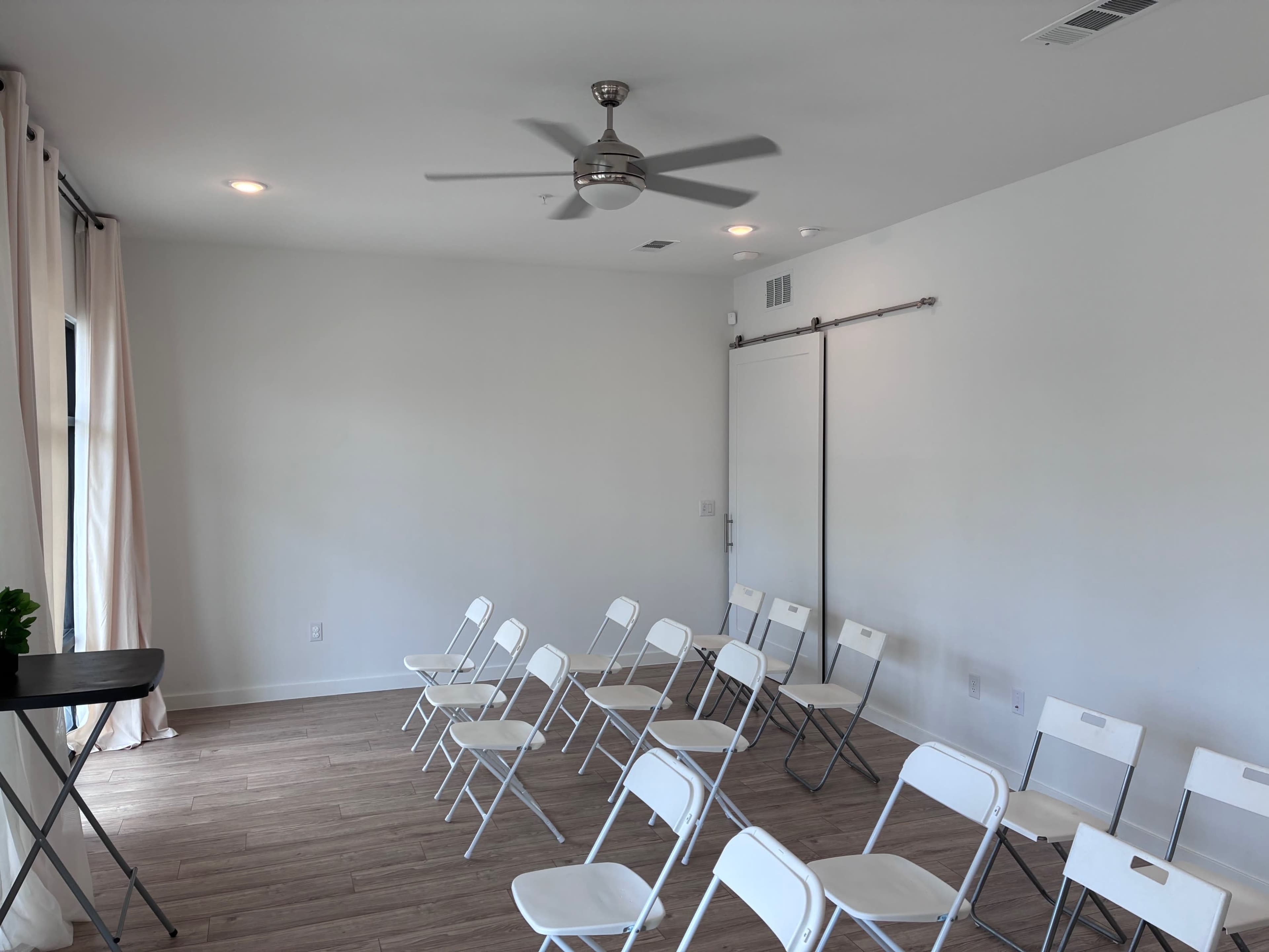 The image shows a room with white walls and wooden flooring, featuring several white folding chairs arranged in rows and a small table in the corner.