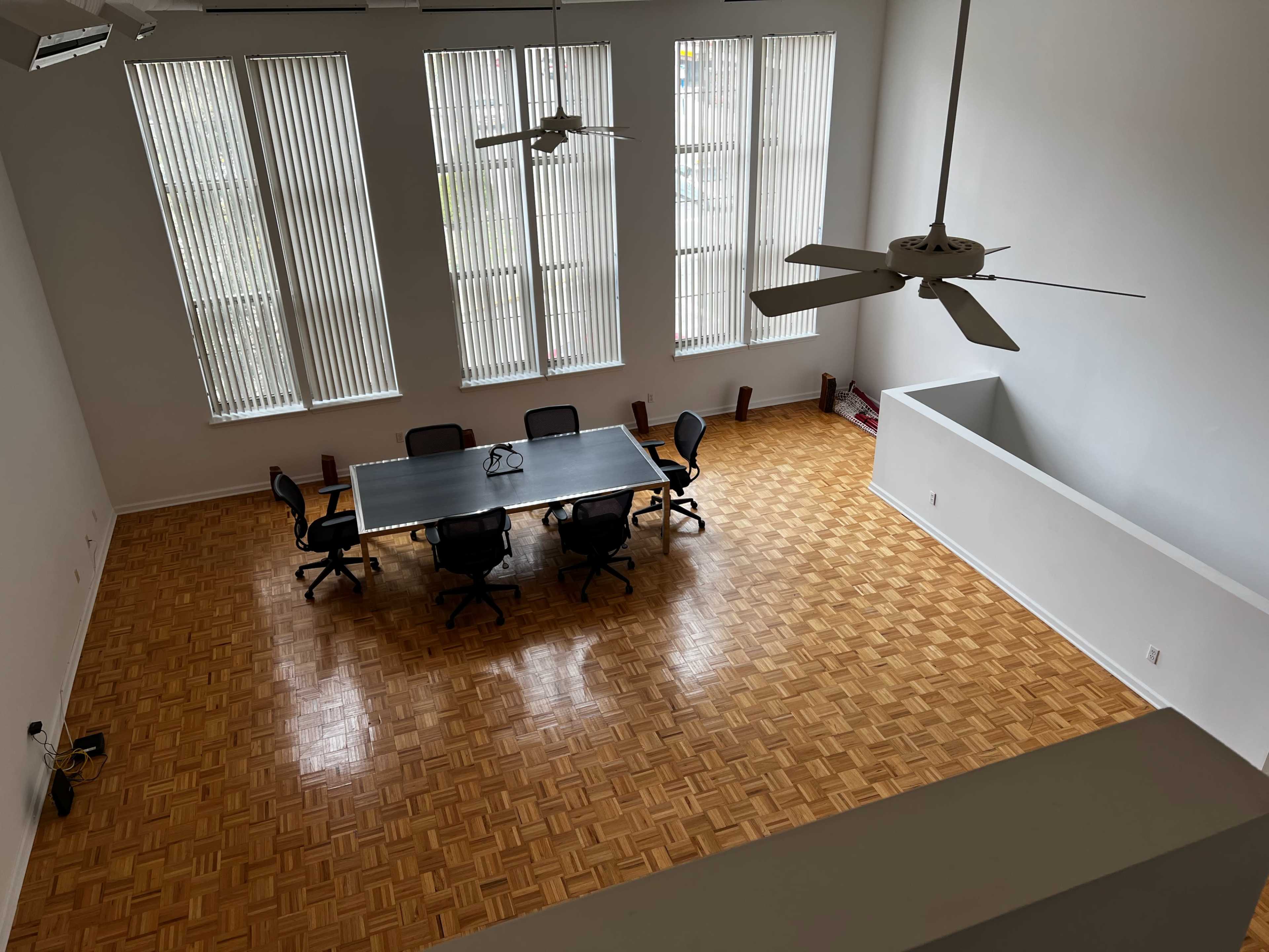 A spacious room with a large table surrounded by chairs, featuring tall windows and a wooden floor.