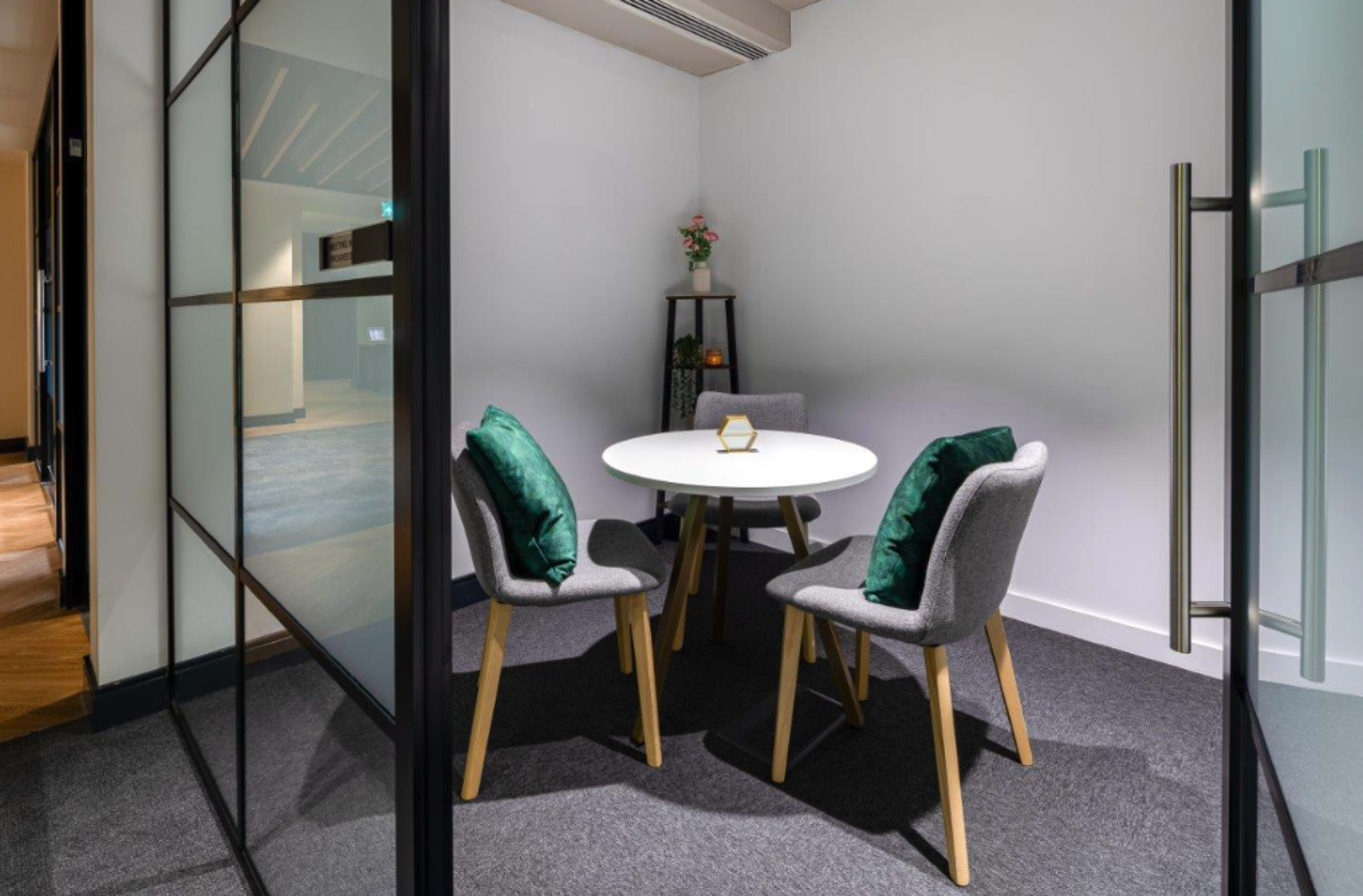 A small meeting room features a round white table surrounded by three gray chairs with green cushions.