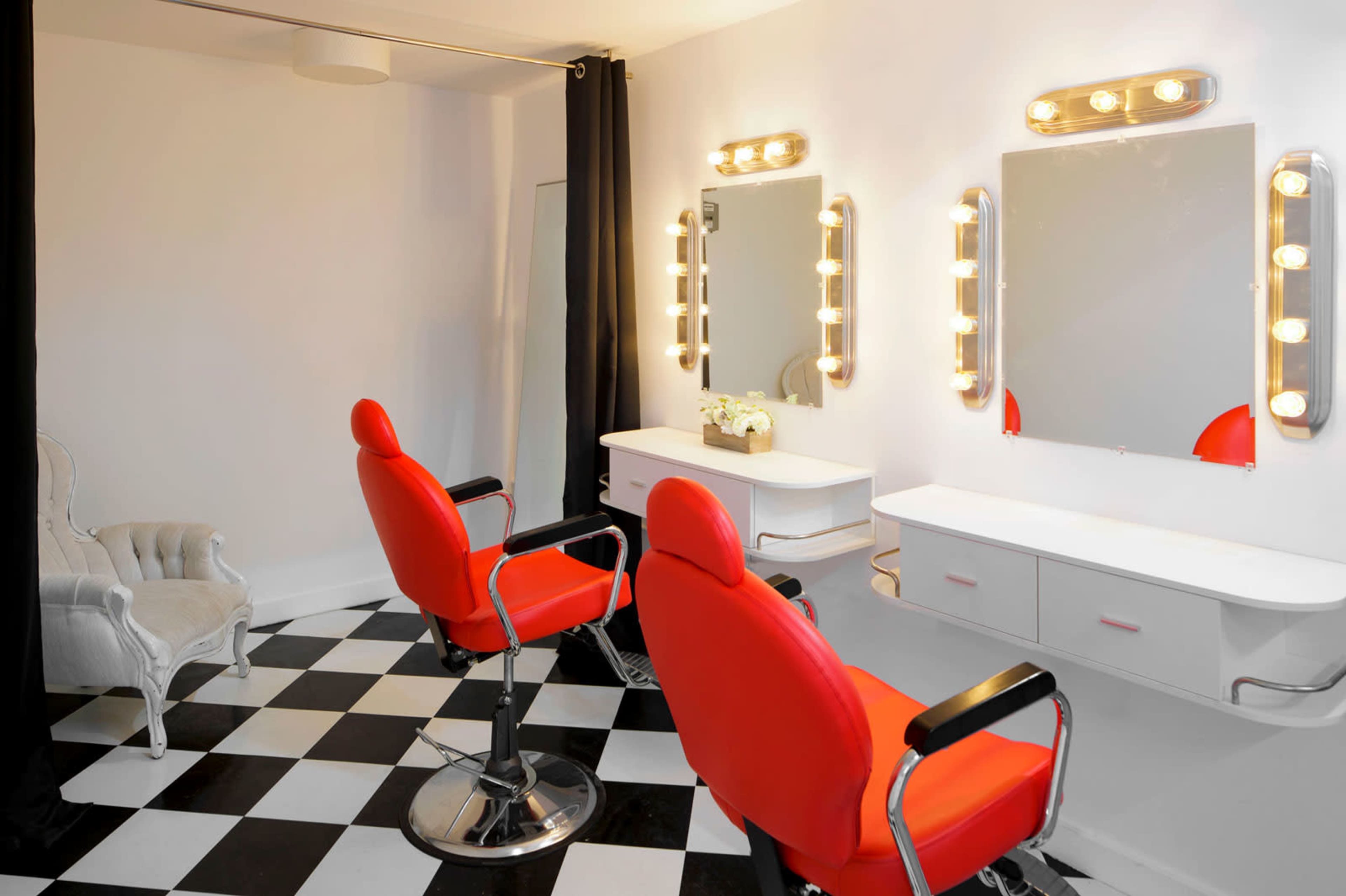 The image shows a minimalist salon interior featuring two red salon chairs in front of mirrors with bright lights, accompanied by a vintage white armchair and a black-and-white checkered floor.