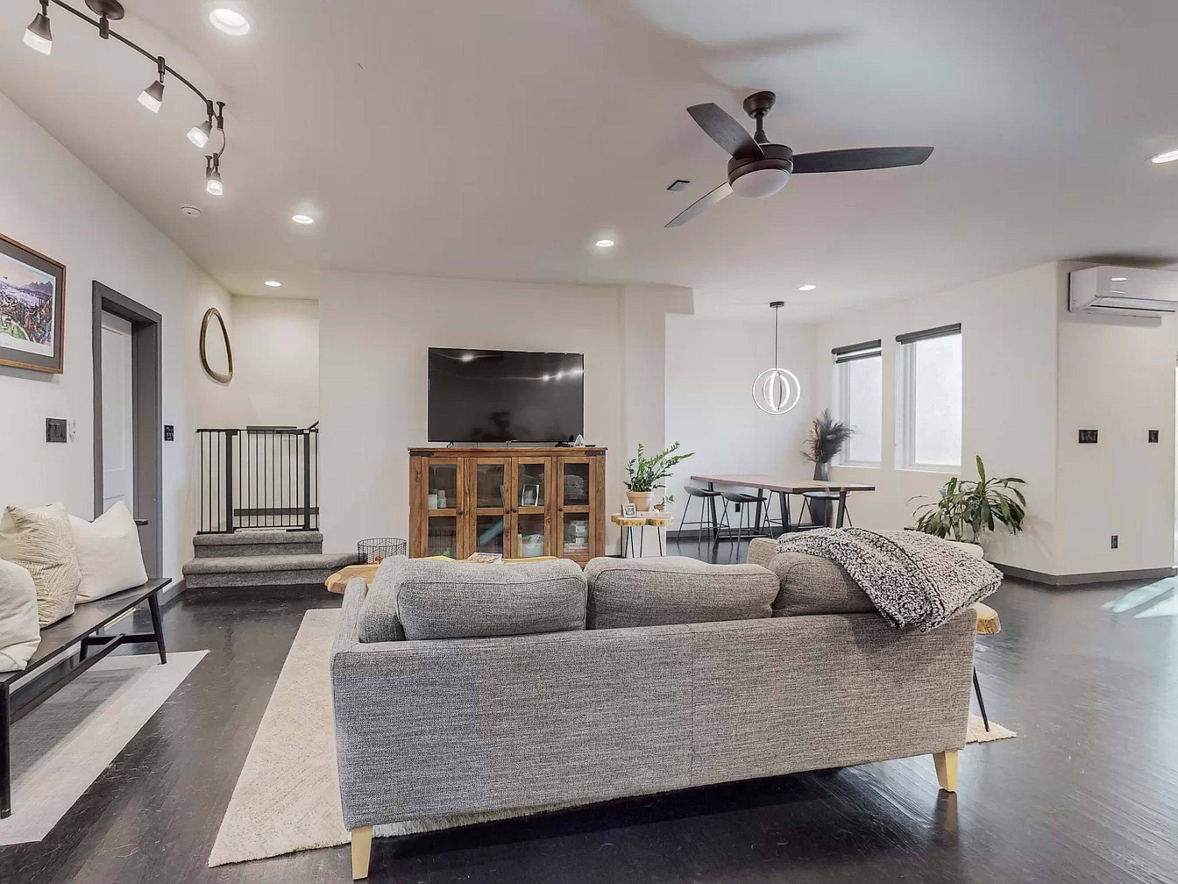 A modern living room with a gray sofa, a wooden TV cabinet, and a dining area in the background featuring a table and plants.