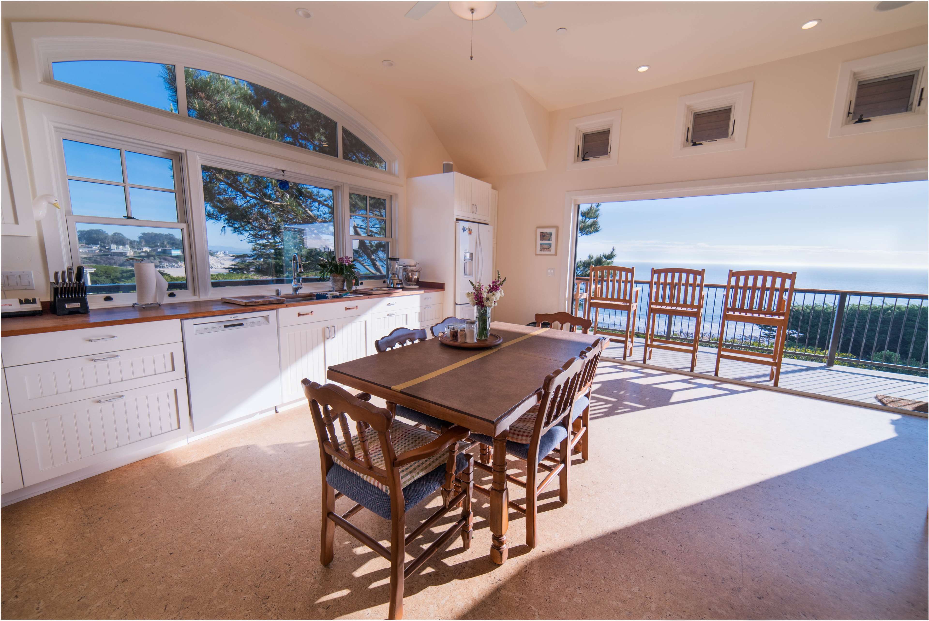 A bright kitchen features a large wooden table surrounded by chairs, with windows offering a view of the outdoors.