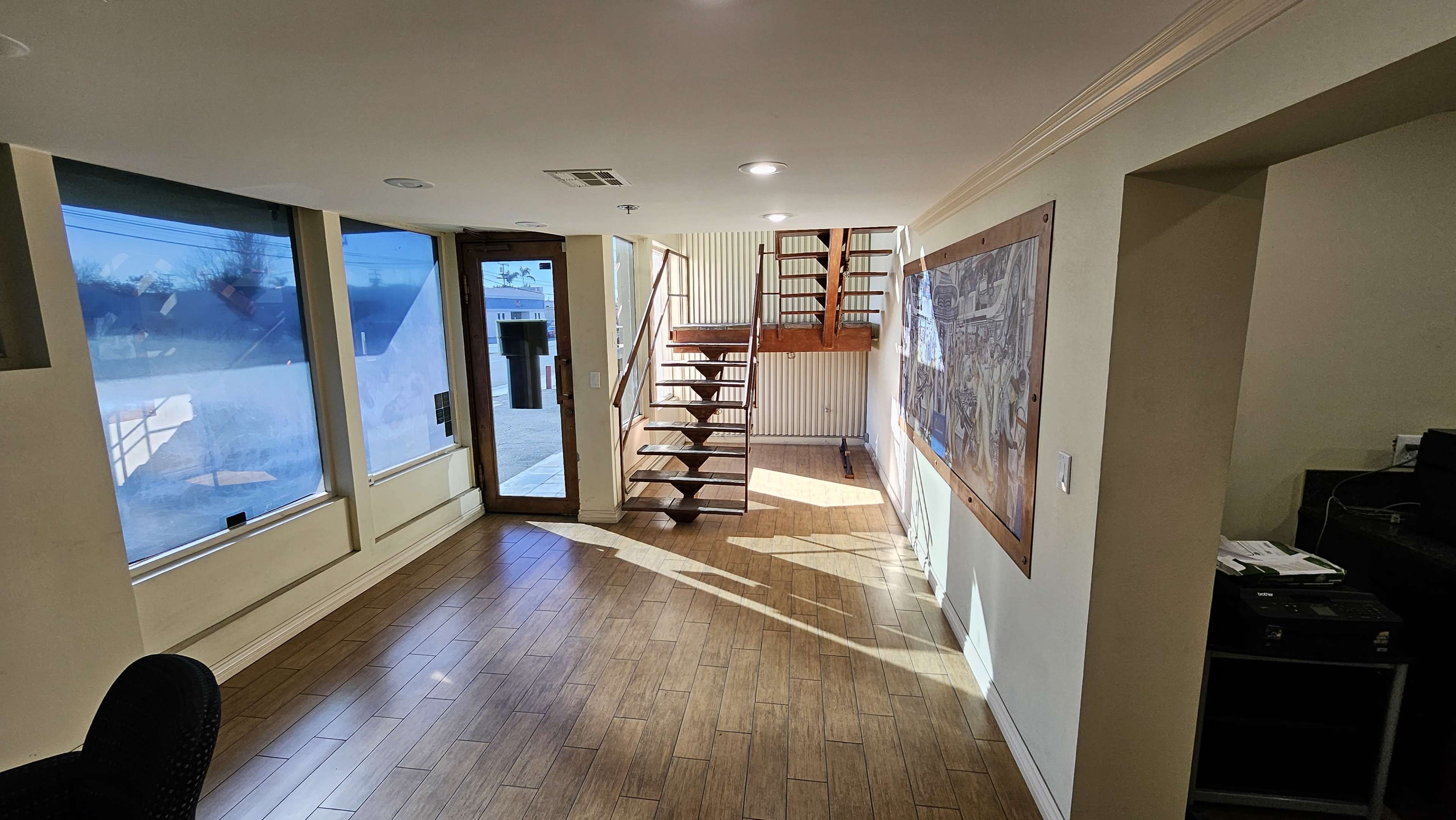 A well-lit interior space with wooden flooring, a staircase leading to an upper level, and large windows allowing natural light to enter.