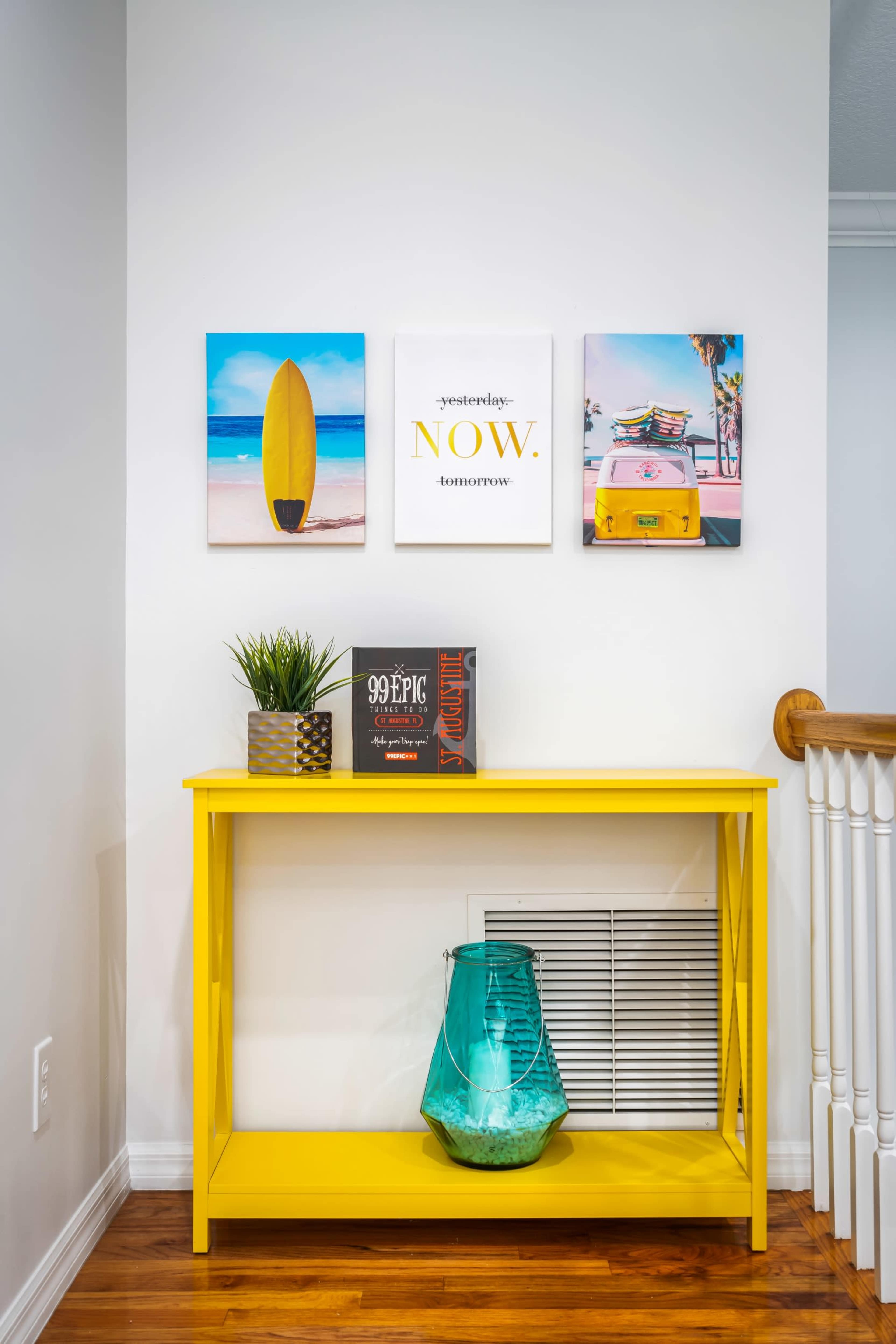 A bright yellow table is placed against a white wall adorned with three colorful framed pictures, complemented by decorative items like a plant and a glass vase.