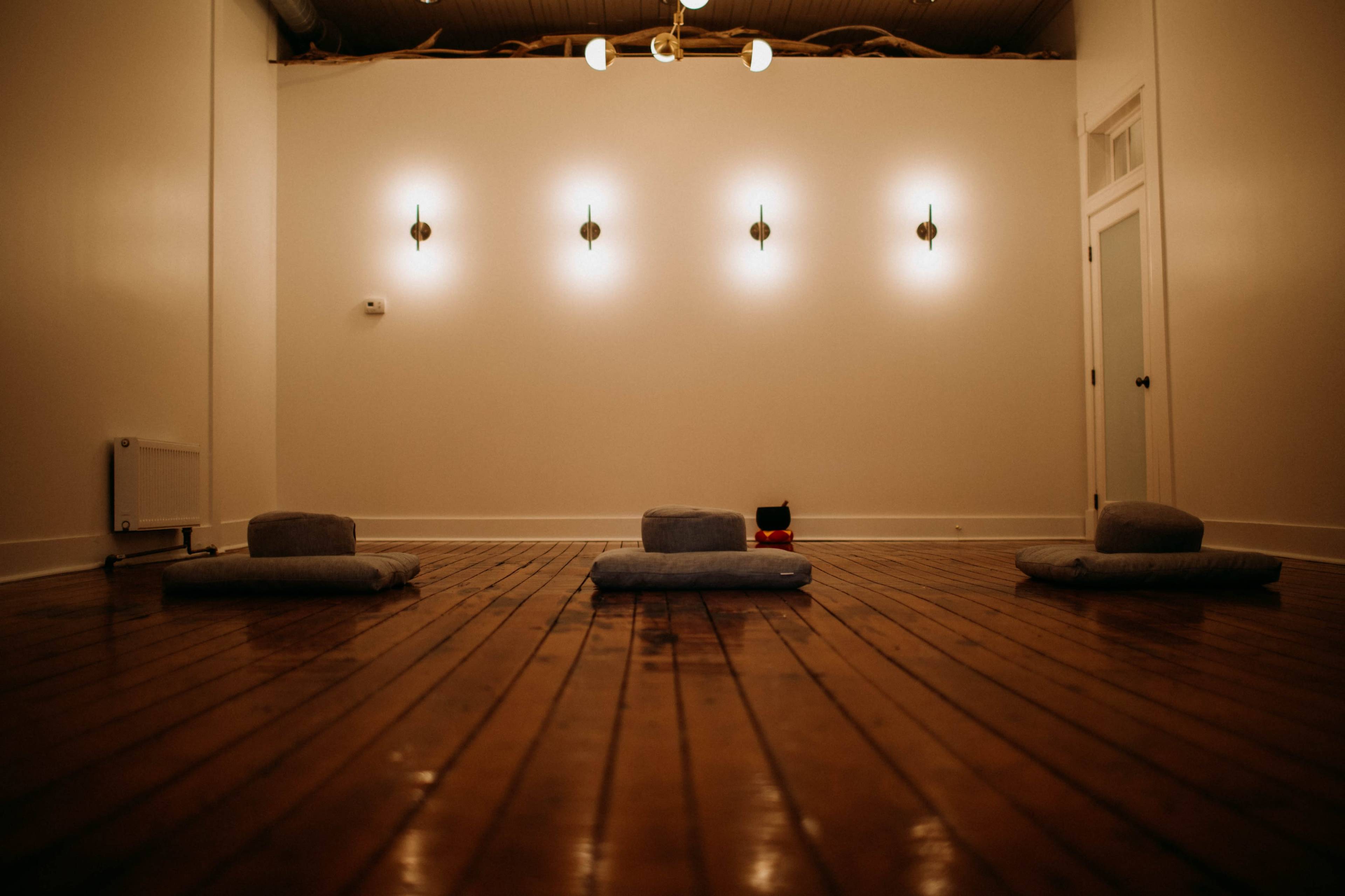 The image shows a minimalist room with wooden flooring, featuring three gray meditation cushions arranged on the floor and several wall-mounted light fixtures.
