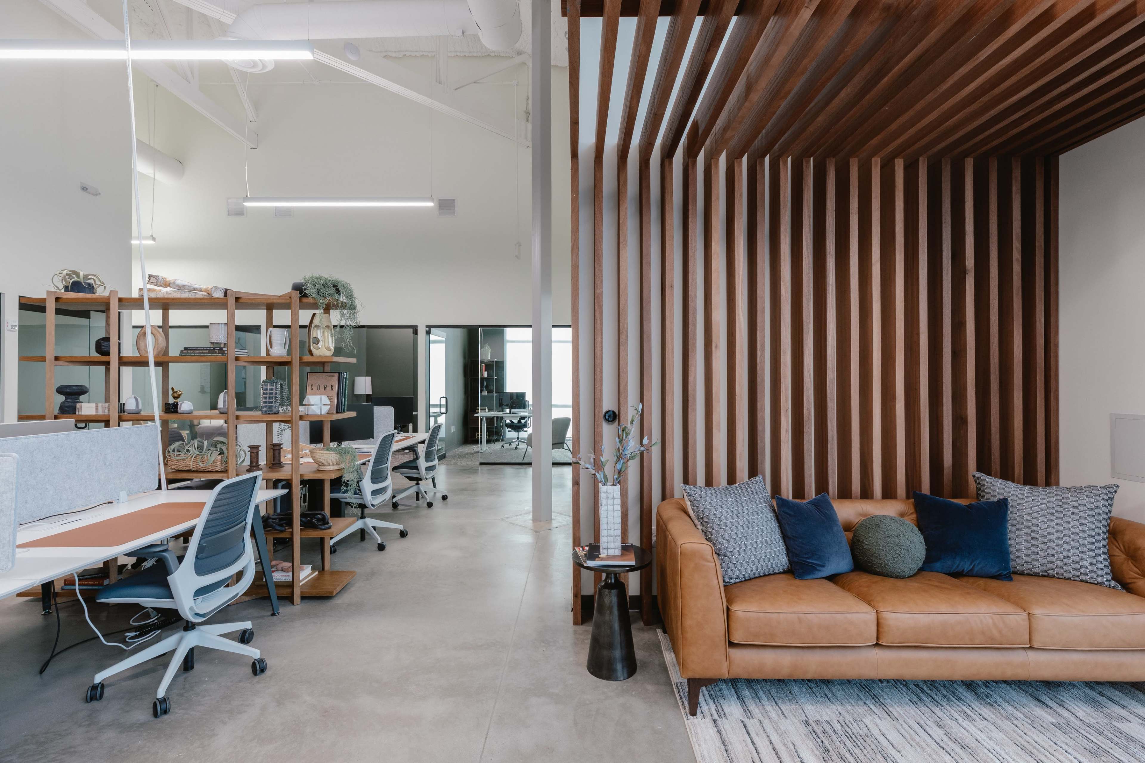 The image shows a modern office space with a wooden slat wall, a brown leather sofa, and multiple workstations with ergonomic chairs.