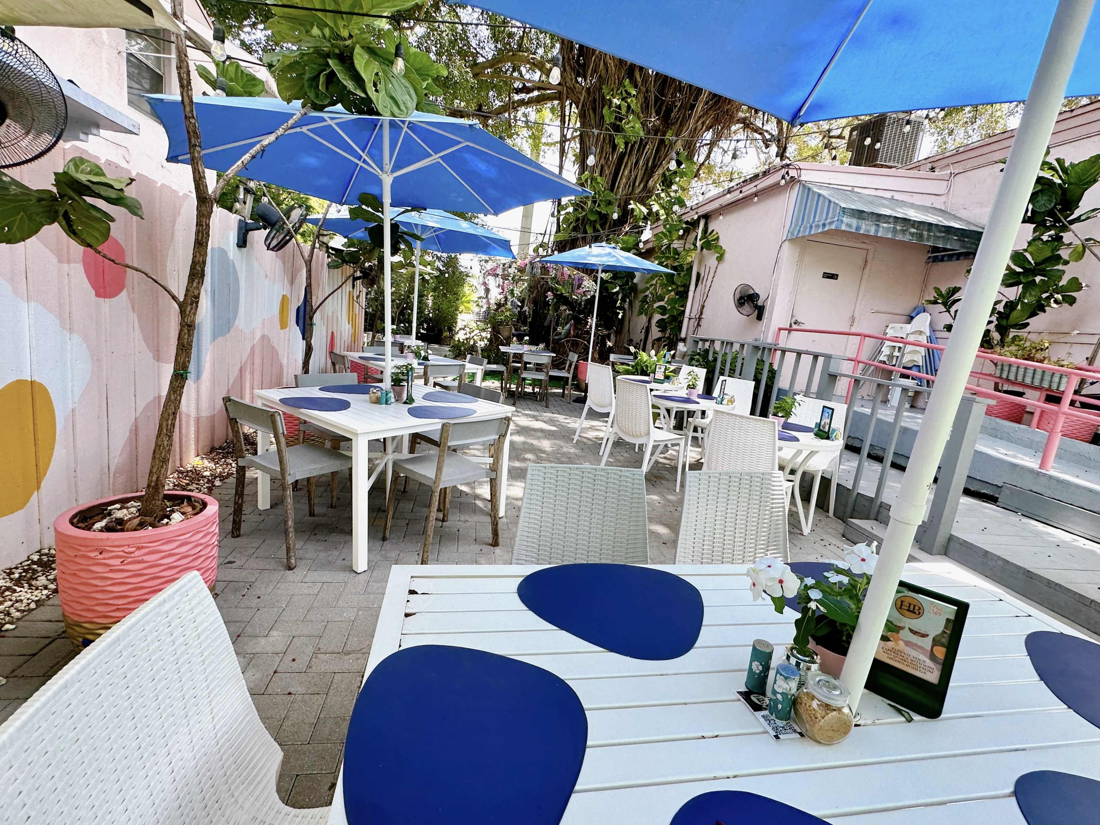A patio dining area features white tables with blue placemats, surrounded by potted plants and umbrellas.