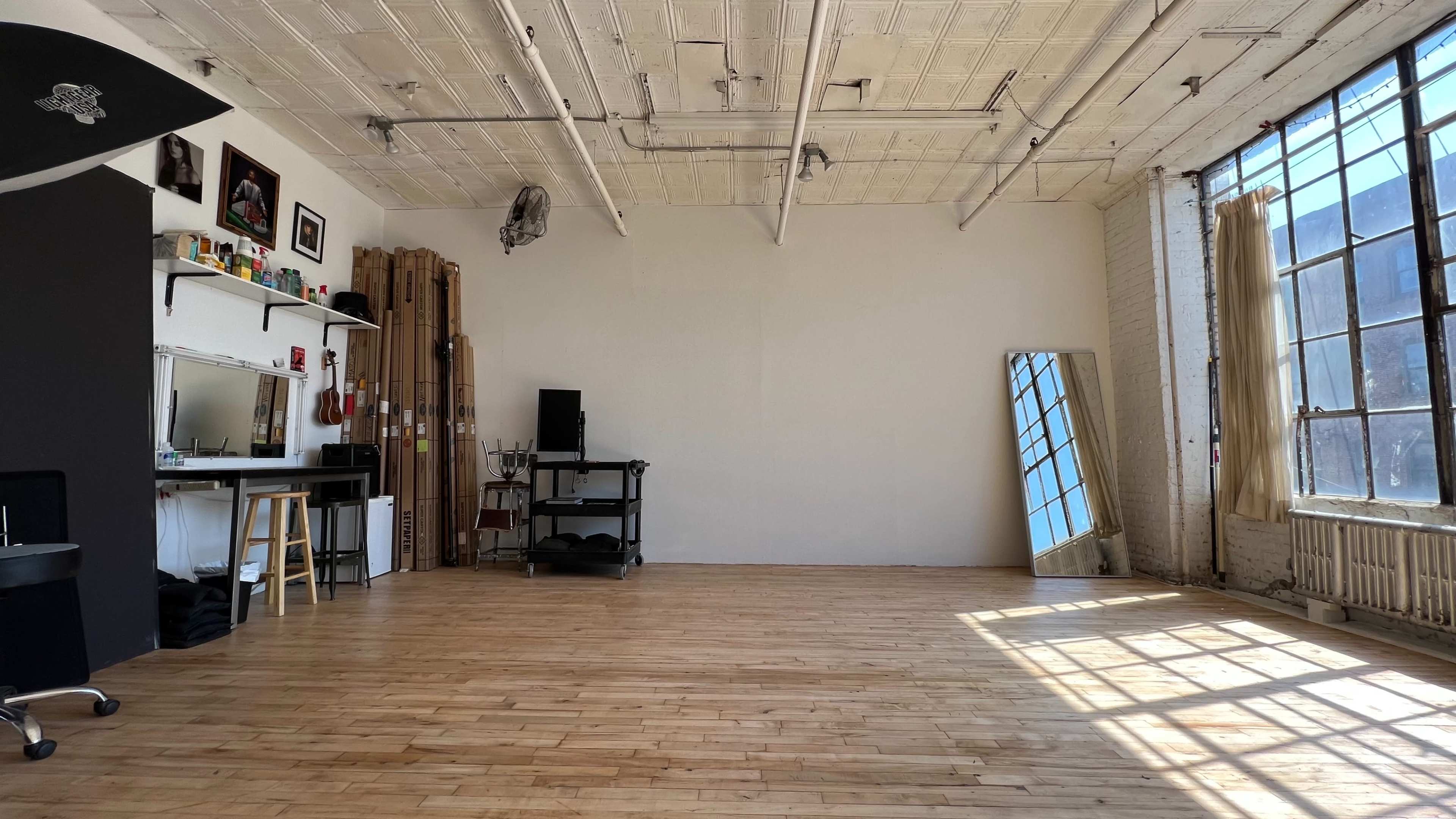 The image shows a spacious, empty room with hardwood floors, large windows, a mirror, and minimal furnishings including a black cart and a stool.