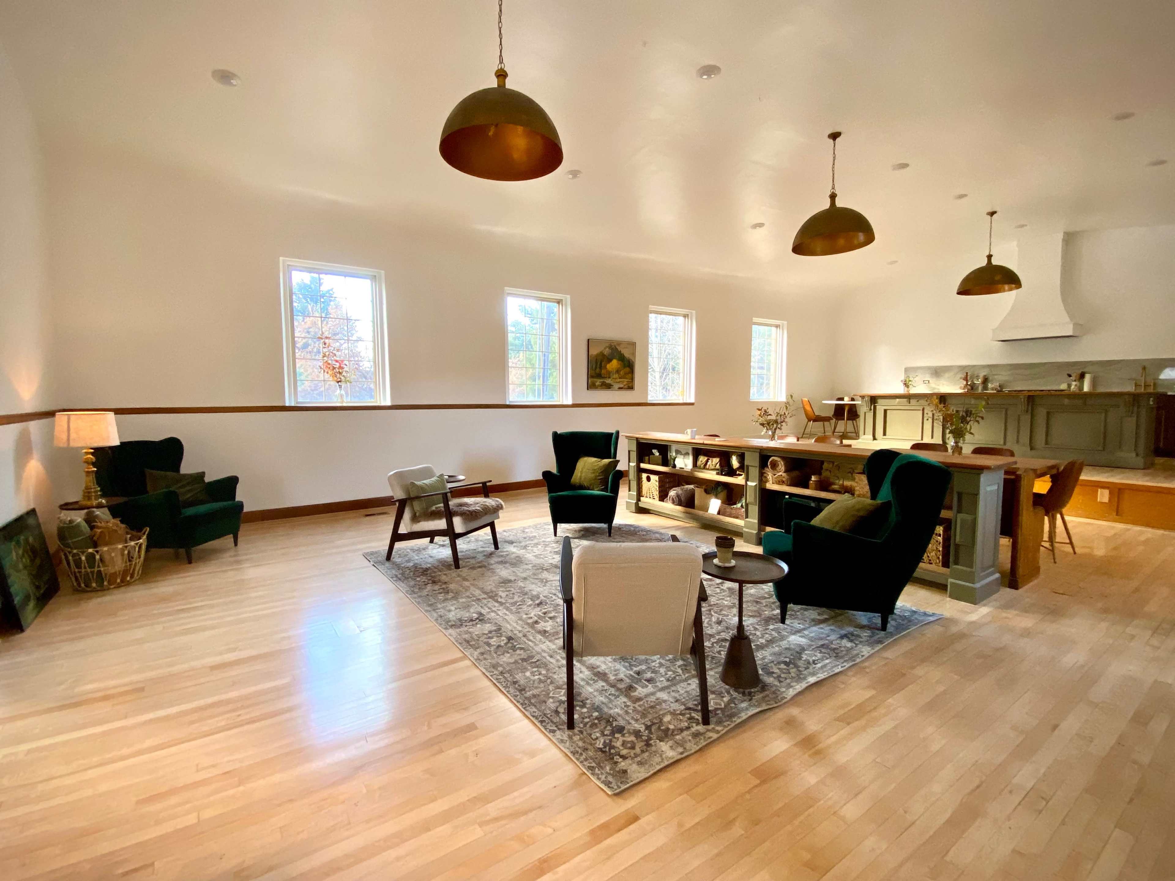 The spacious room features green chairs, a coffee table, a rug, and large windows that bring in natural light.