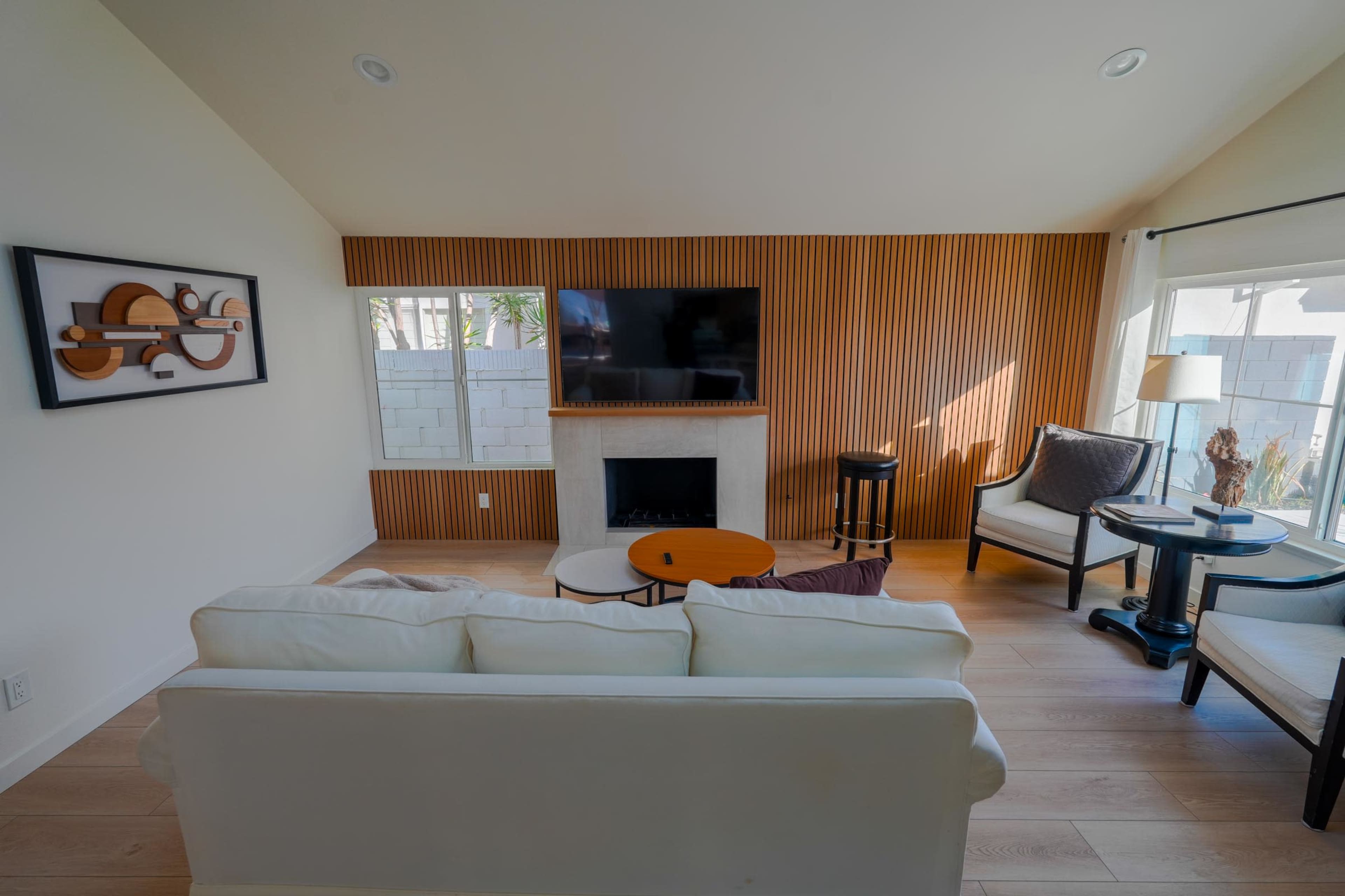 The image shows a modern living room with a white sofa, a wooden accent wall behind a television, and a small coffee table in front of a fireplace.