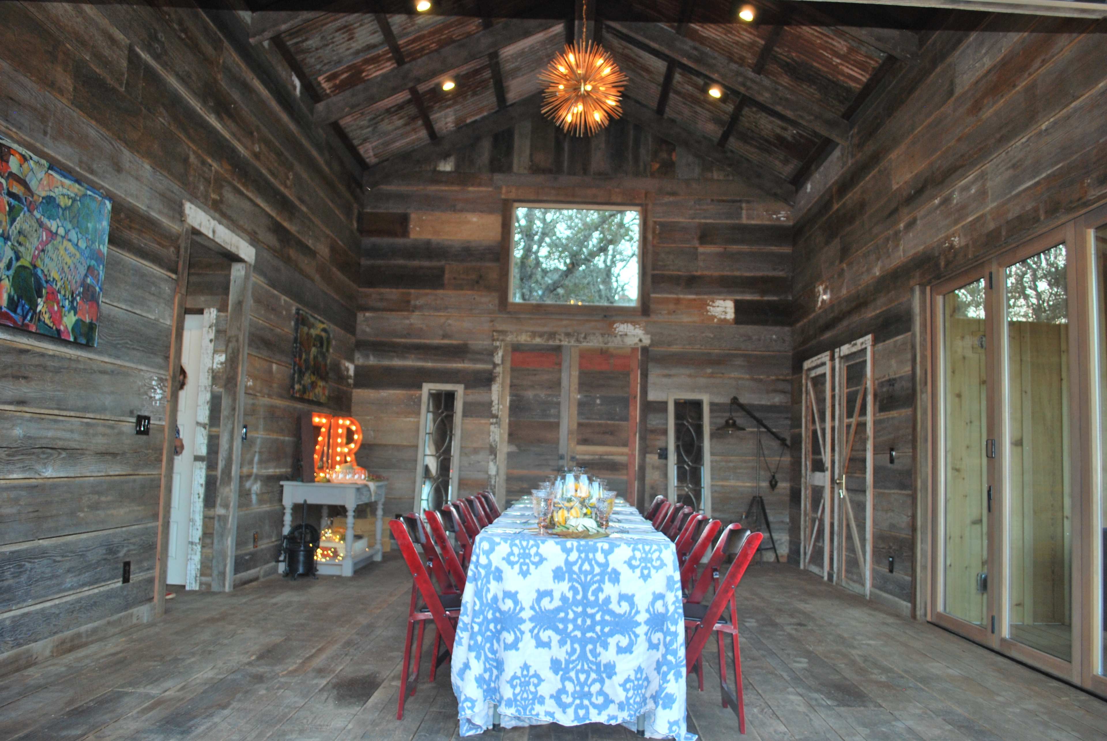 The interior of a rustic dining room features a long table set with a blue tablecloth and red chairs, surrounded by weathered wooden walls and large windows.