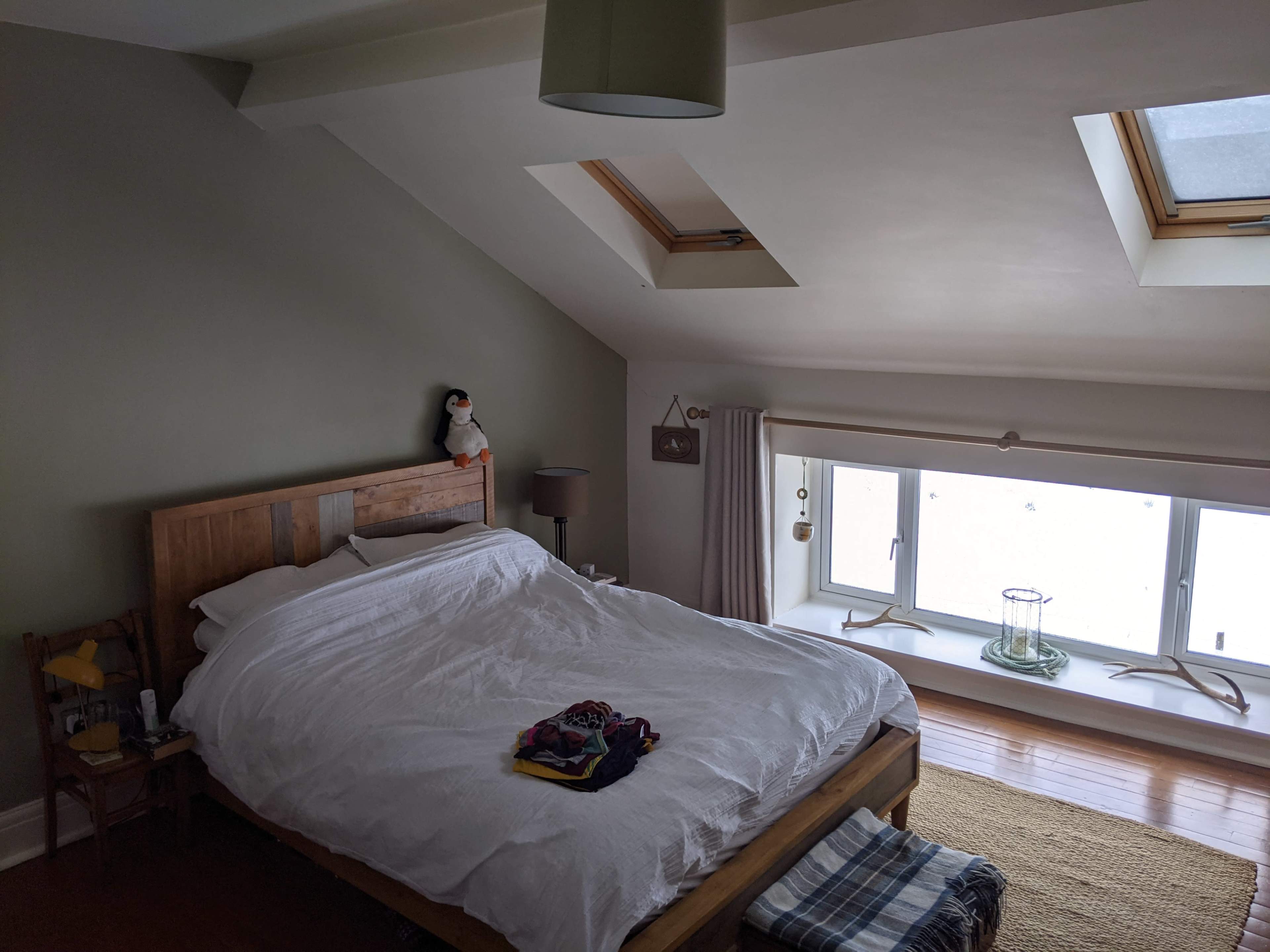 A bedroom features a large bed with white bedding, a penguin plush toy on the headboard, and a window with skylights above.
