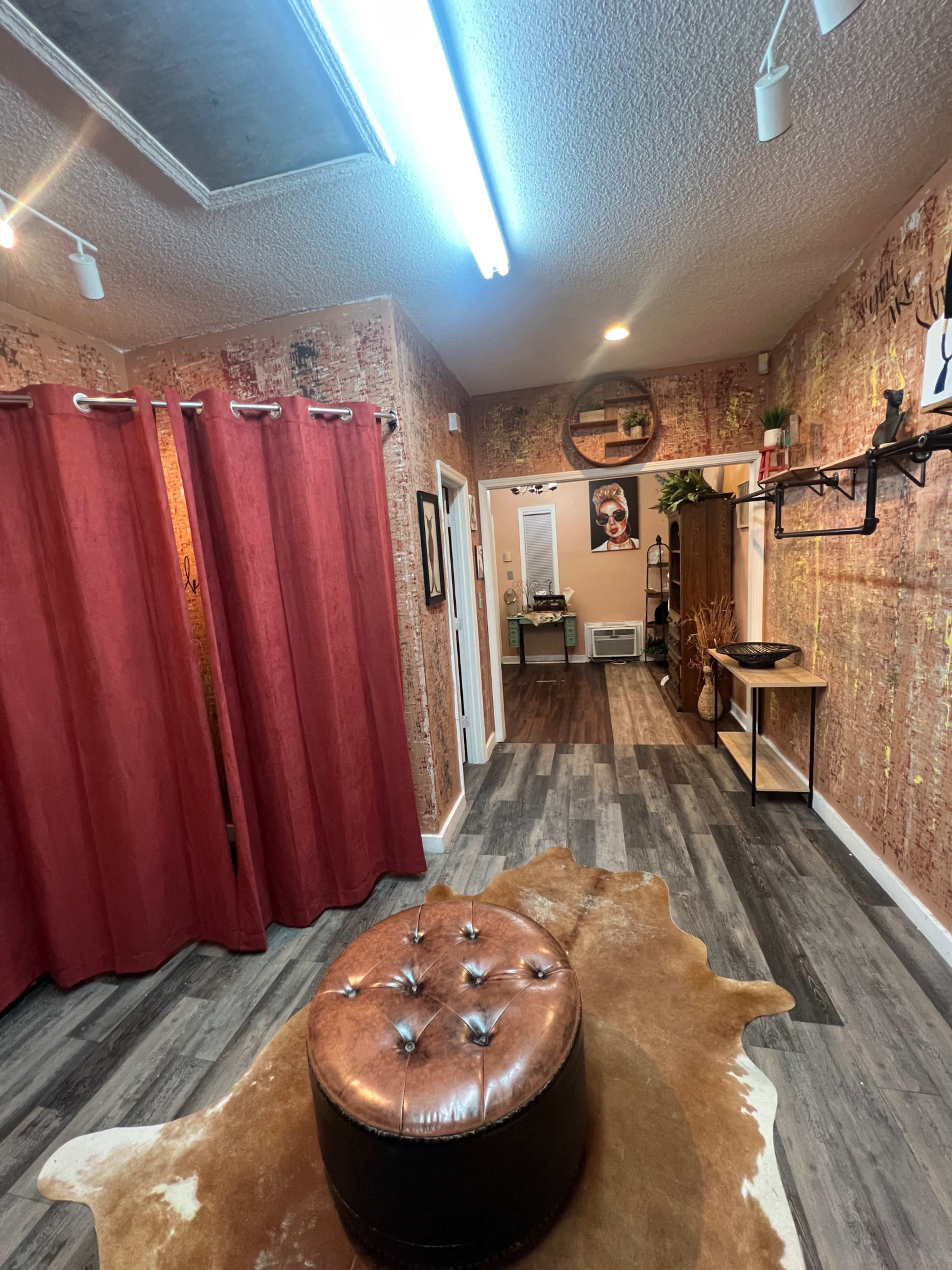 The image shows a room with red curtains, wood flooring, and a decorative circular mirror on the wall, featuring a brown leather ottoman in the center.