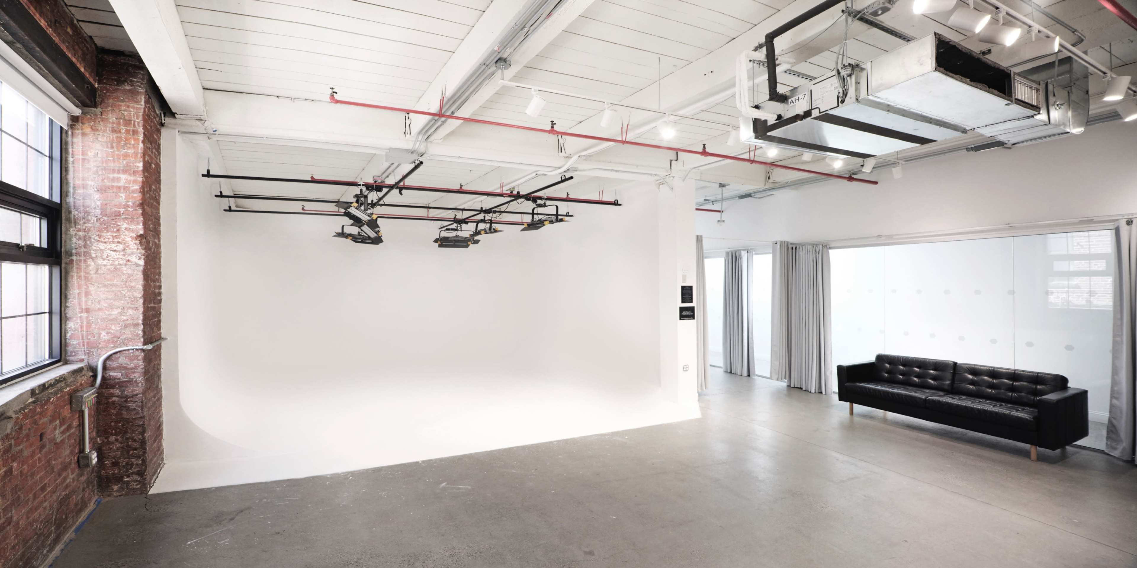 A spacious studio with a white backdrop, hanging lights, exposed brick walls, and a black leather couch in one corner.