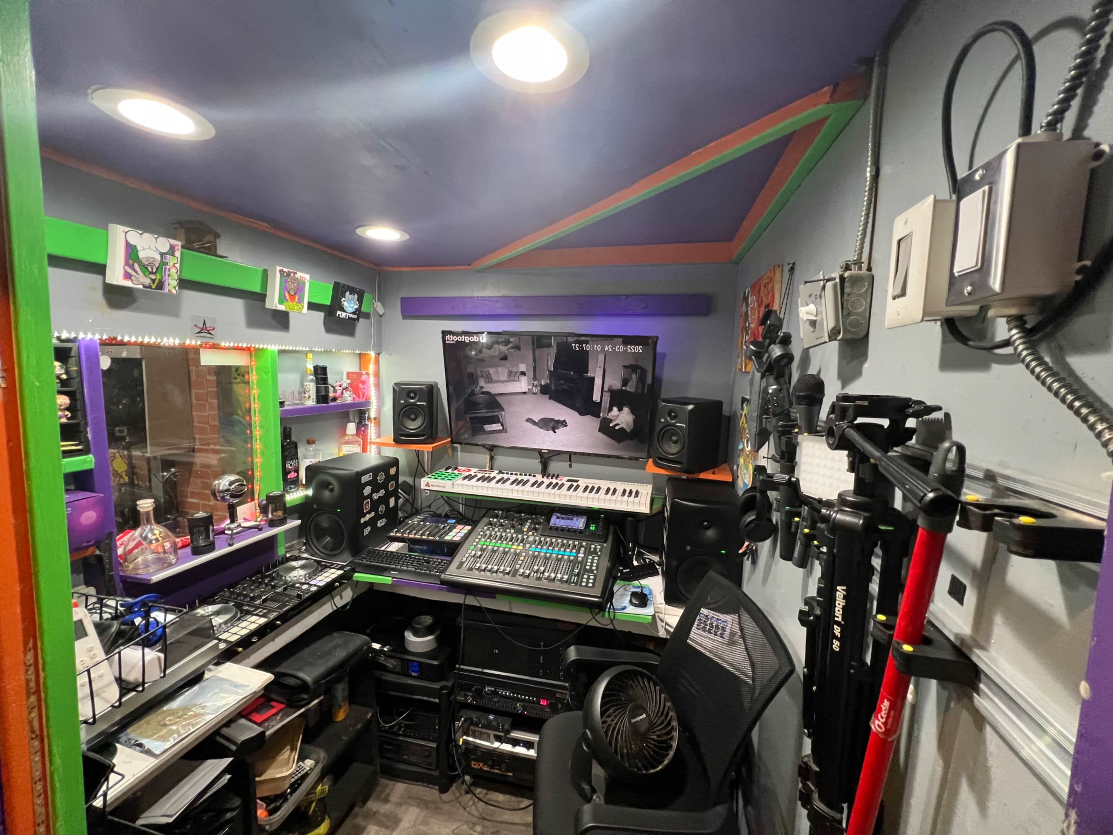 A compact music studio filled with audio equipment, a large screen displaying a camera feed, and colorful decorations on the walls.
