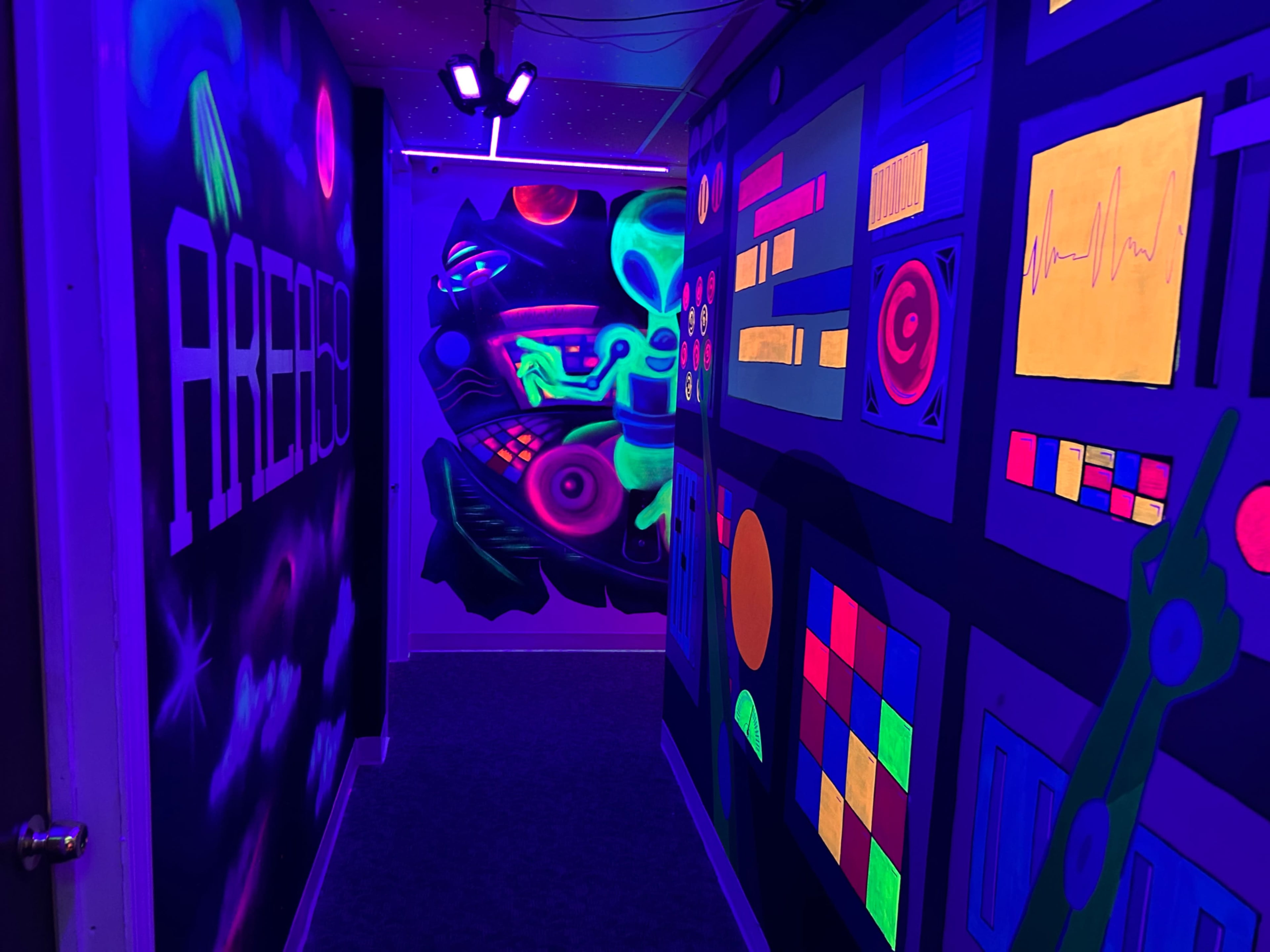 A colorful hallway illuminated by black lights, featuring vibrant murals on the walls depicting abstract shapes and futuristic designs.
