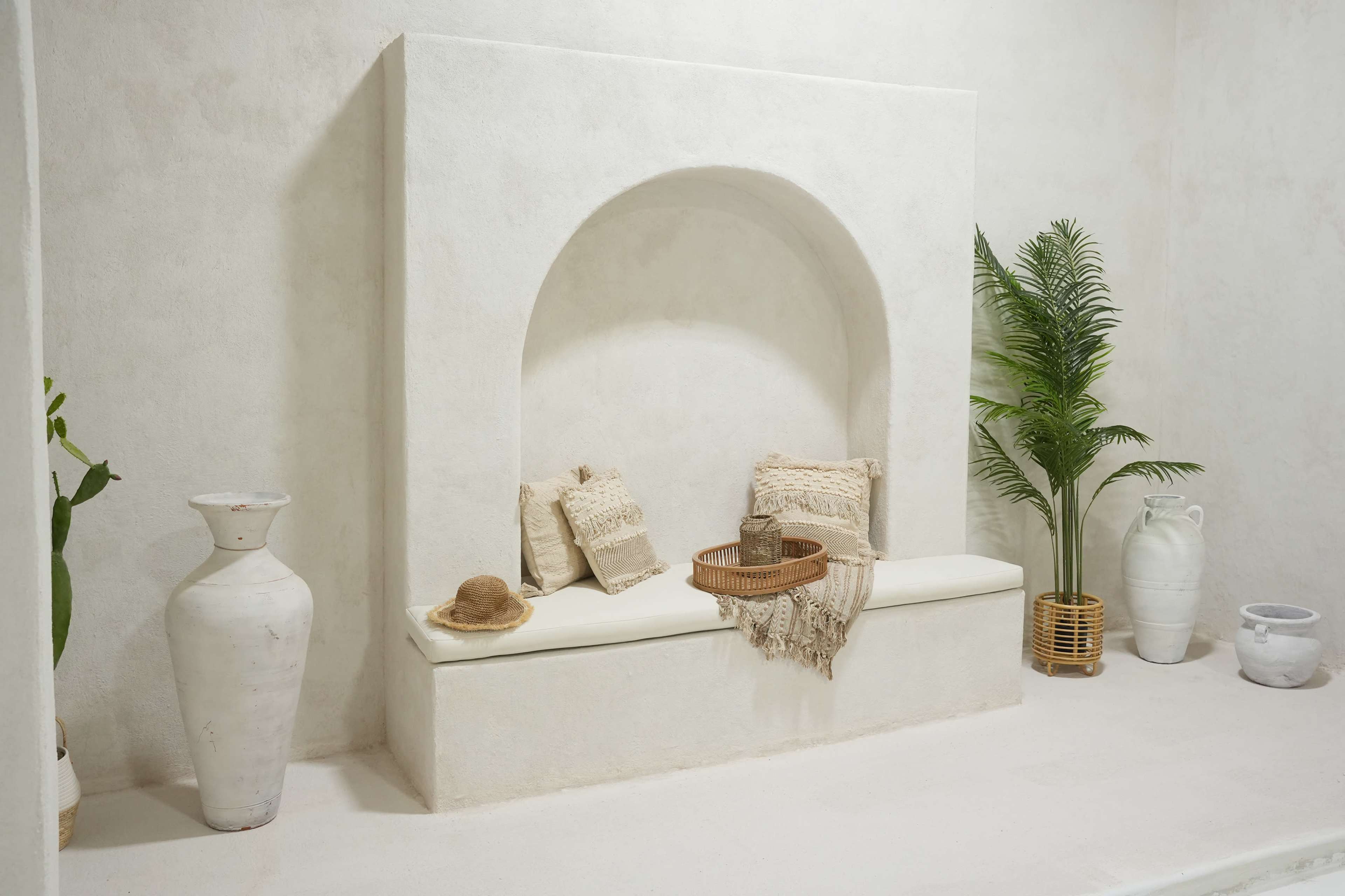 A minimalist interior with a white stucco wall featuring a curved alcove bench adorned with textured cushions, surrounded by decorative plants and pottery.